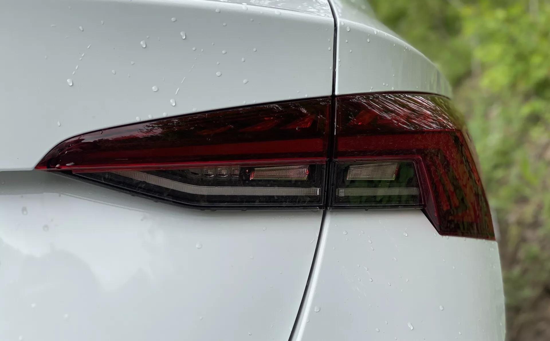 Vue rapprochée du côté arrière gauche d'une Škoda Octavia 2020, mettant en avant l'ensemble de feux arrière. L'image est prise sous un angle diagonal soulignant le design contemporain du feu arrière à LED avec des angles nets et une finition brillante. Une partie du panneau arrière et du couvercle de coffre sont visibles, montrant une intégration harmonieuse et une esthétique aérodynamique. La surface lisse présente des gouttes d'eau, indiquant des précipitations récentes ou un lavage récent.