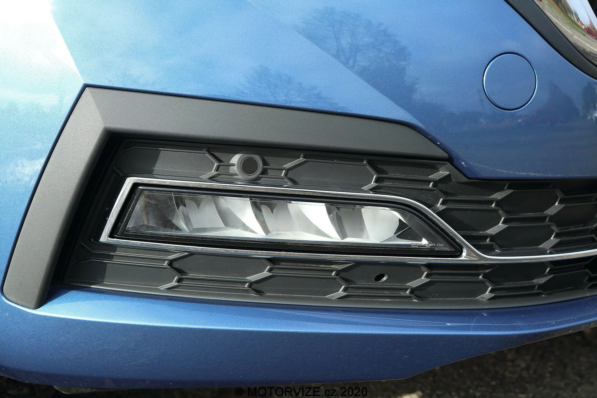 Détail rapproché de l'avant de la Skoda Octavia 2020 (Octavia Limousine), mettant en avant le phare gauche et la grille. Le phare LED moderne avec un design angulaire et aigu est placé dans un renfoncement sculpté, fusionnant parfaitement avec la carrosserie bleue métallisée de la voiture. La grille adjacente présente un motif hexagonal en plastique noir avec des accents chromés, reflétant le style automobile contemporain.