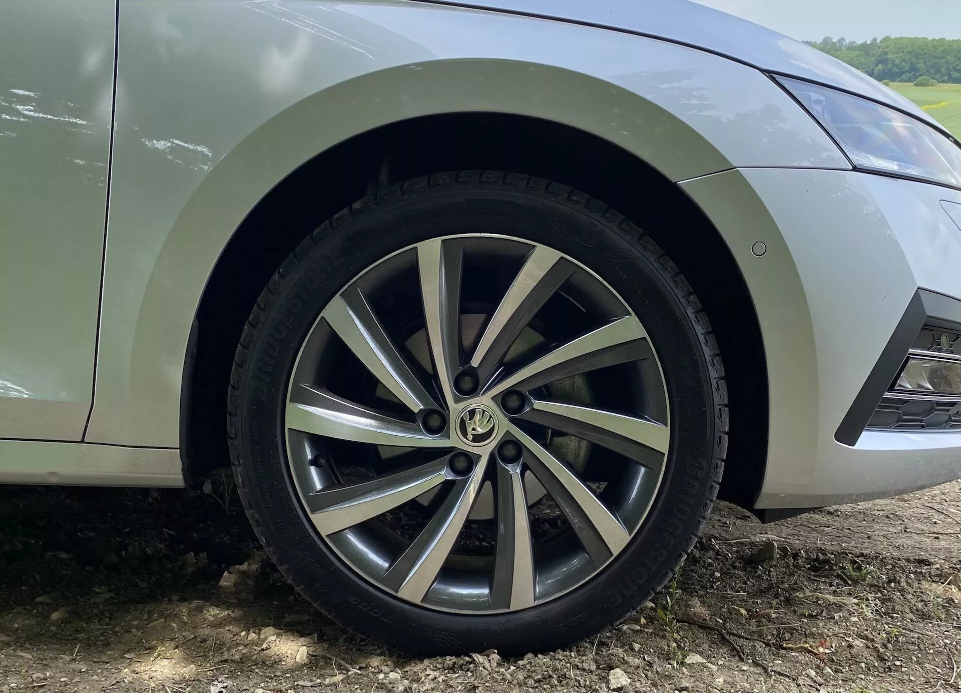 Gros plan sur la roue avant et l'aile inférieure d'une Skoda Octavia Limousine 2020, mettant en valeur le design de la jante en alliage à rayons multiples avec un mélange de surfaces peintes en argent et usinées. La bande de roulement du pneu est visible et montre peu d'usure. La voiture est garée sur une surface en terre avec des éclaboussures de boue visibles derrière la roue, indiquant une utilisation récente hors route ou sur une route non pavée. Seule la section de la voiture allant de la portière avant inférieure au pare-chocs avant est montrée sous un angle bas, capturant les détails de la roue et de l'aile.