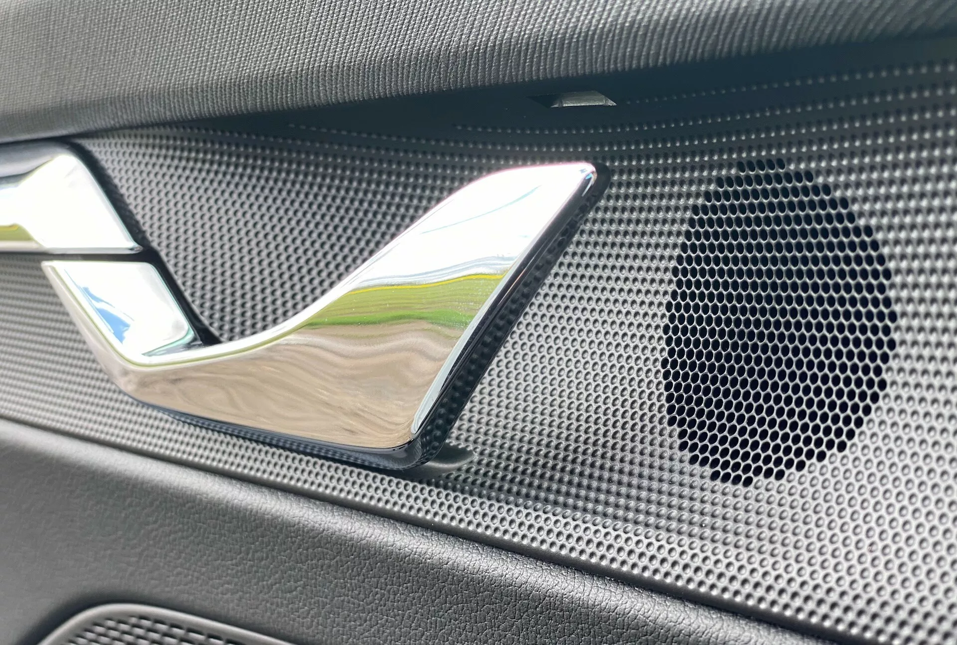 Image en gros plan de l'intérieur de la Skoda Octavia limousine 2020, mettant en avant la poignée de porte chromée et la grille du haut-parleur avec motif en nid d'abeille dans un matériau texturé, peut-être en fibre de carbone, soulignant le luxe et le système audio haute-fidélité.