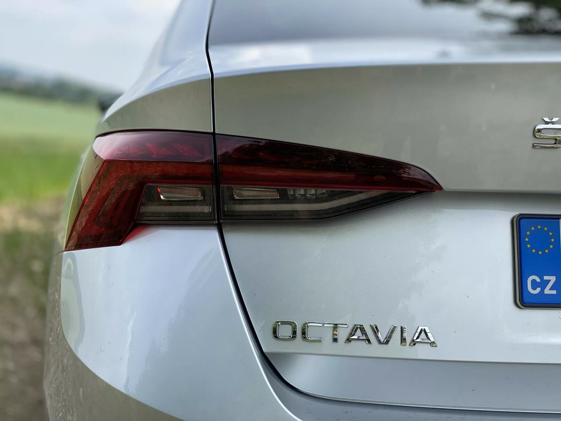 Vue en gros plan de la section arrière droite d'une Škoda Octavia 2020 de couleur argentée. L'image capture le design net et angulaire du feu arrière avec des éléments intégrés pour les feux de freinage, les clignotants et peut-être les feux de recul. Le coffre affiche en bonne place la désignation du modèle 'OCTAVIA' en lettres chromées sous le feu arrière. Un petit rectangle bleu et jaune avec des étoiles indique l'immatriculation de la République tchèque. La carrosserie du véhicule, avec une finition métallique et polie, reflète l'environnement ambiant, avec un arrière-plan de verdure flou qui met en valeur son design sophistiqué.