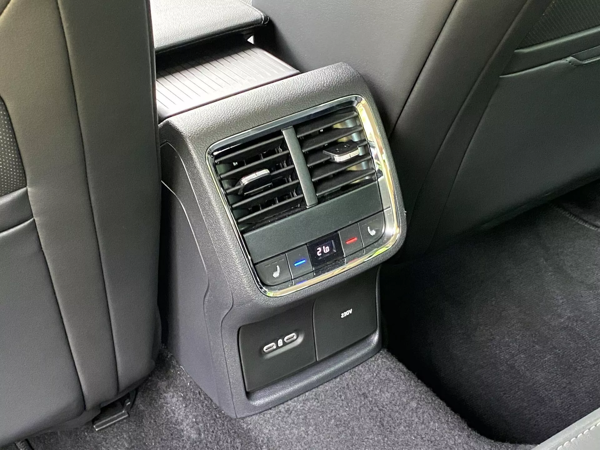 Vue détaillée de la console de commande du climat arrière à l'arrière d'une Skoda Octavia limousine 2020. Capturée sous un angle décentré, offrant une vue claire du panneau central situé à l'arrière de l'accoudoir avant. Comprend des boutons de contrôle climatique numérique avec des réglages de température, des aérateurs verticaux, un affichage numérique de la température réglé à 21℃, des ports de chargement USB et une fonctionnalité potentielle de chauffage ou de refroidissement des sièges arrière. Les matériaux sont un mélange de plastique et de tissu, dans un schéma de couleurs noir et gris foncé sous éclairage naturel.
