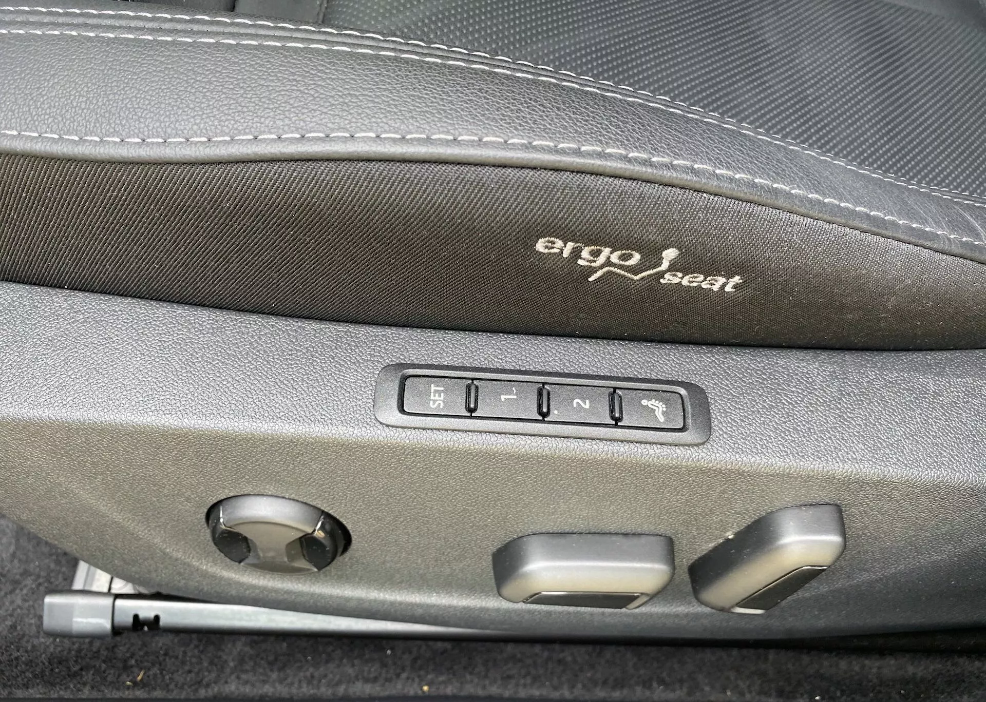 Vue rapprochée de l'intérieur de la Škoda Octavia 2020, mettant en avant les commandes de réglage ergonomique du siège. 'Siège ergo' est brodé sur la partie supérieure sombre du siège. Sous la broderie, cinq boutons sont visibles : un pour les réglages de mémoire du siège et quatre pour les ajustements du siège comme l'avance, le recul, la montée, la descente et l'inclinaison du dossier. Un bouton rond est également présent, probablement pour ajuster le soutien lombaire ou d'autres réglages multifonctionnels du siège, indiquant un confort supérieur et une technologie avancée dans ce véhicule.