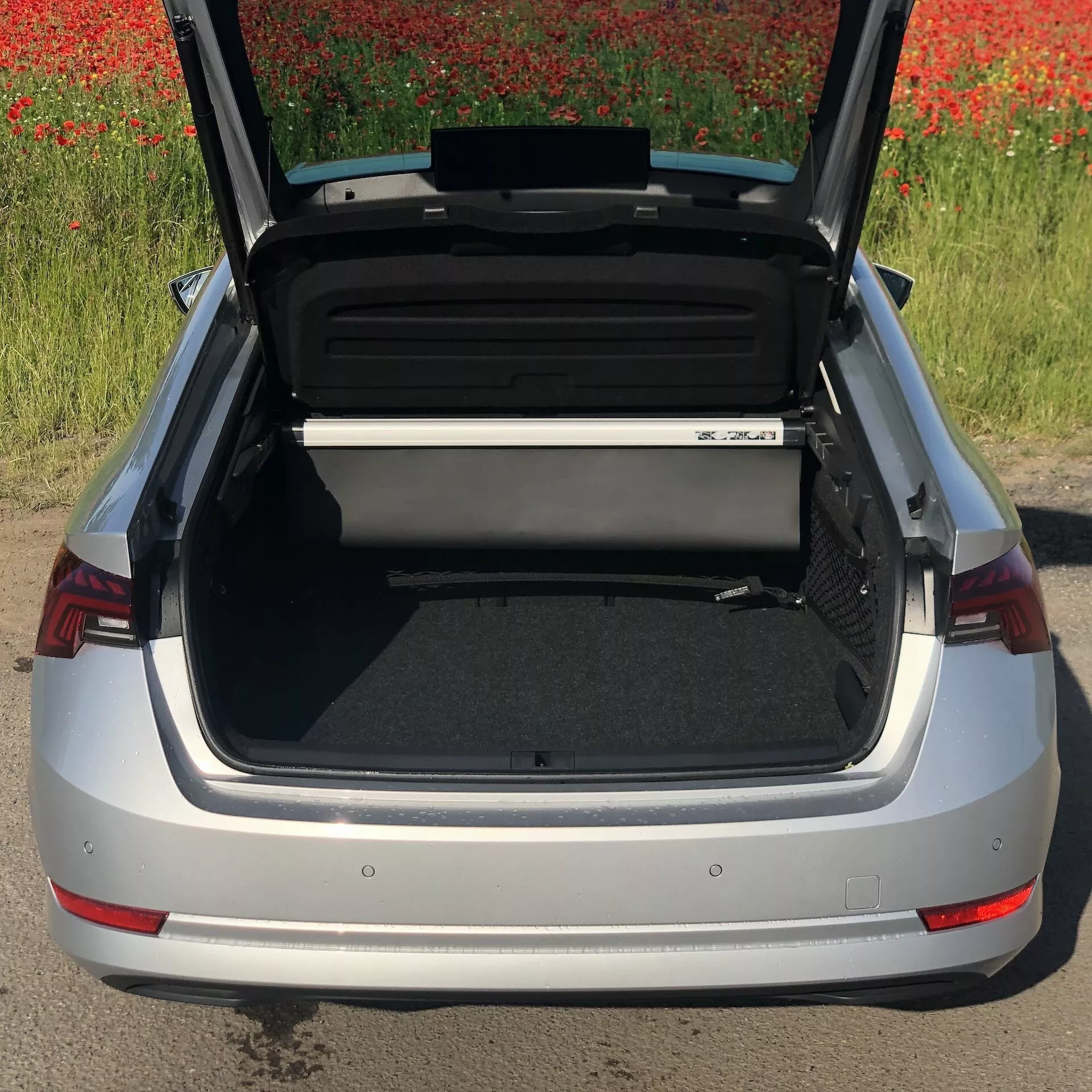 Vue en plongée d'une Skoda Octavia 2020 argentée avec son coffre ouvert, montrant l'espace de chargement spacieux et organisé. Le pare-chocs arrière, les feux arrière distinctifs rouges et une partie de l'emblème de la voiture sont visibles, avec un champ de fleurs rouges en arrière-plan.