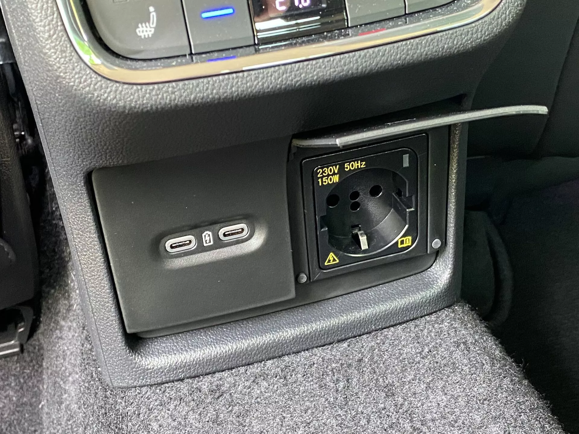Vue rapprochée des caractéristiques de l'alimentation électrique de l'intérieur de la Skoda Octavia 2020 : partie inférieure de la console centrale avec deux ports USB-A à gauche pour la recharge ou la connexion multimédia et une prise secteur 230V AC pour fiches européennes jusqu'à 150W à droite, entourée d'une finition texturée et d'une moquette visible.
