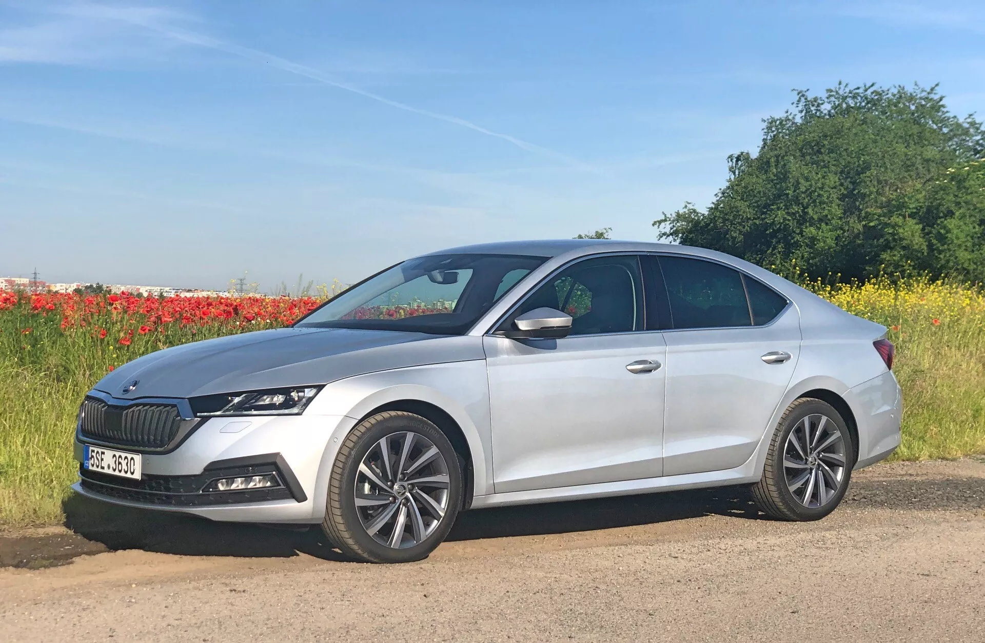 L'image montre une berline argentée à quatre portes, spécifiquement la Skoda Octavia 2020 (Octavia-Limousine), garée à côté d'une route avec un arrière-plan naturel comprenant un champ et une ligne d'arbres. Photografiée sous un angle de profil latéral légèrement avancé, elle présente des roues en alliage à plusieurs rayons, des phares LED angulaires élégants et une calandre proéminente avec des lamelles verticales. Le design moderne comprend des lignes sculptées sur la carrosserie et une ligne de toit inclinée, avec des rétroviseurs latéraux intégrant des clignotants. D'autres caractéristiques visibles incluent une antenne arrière et des capteurs sur le pare-chocs avant indiquant des systèmes avancés d'assistance à la conduite.