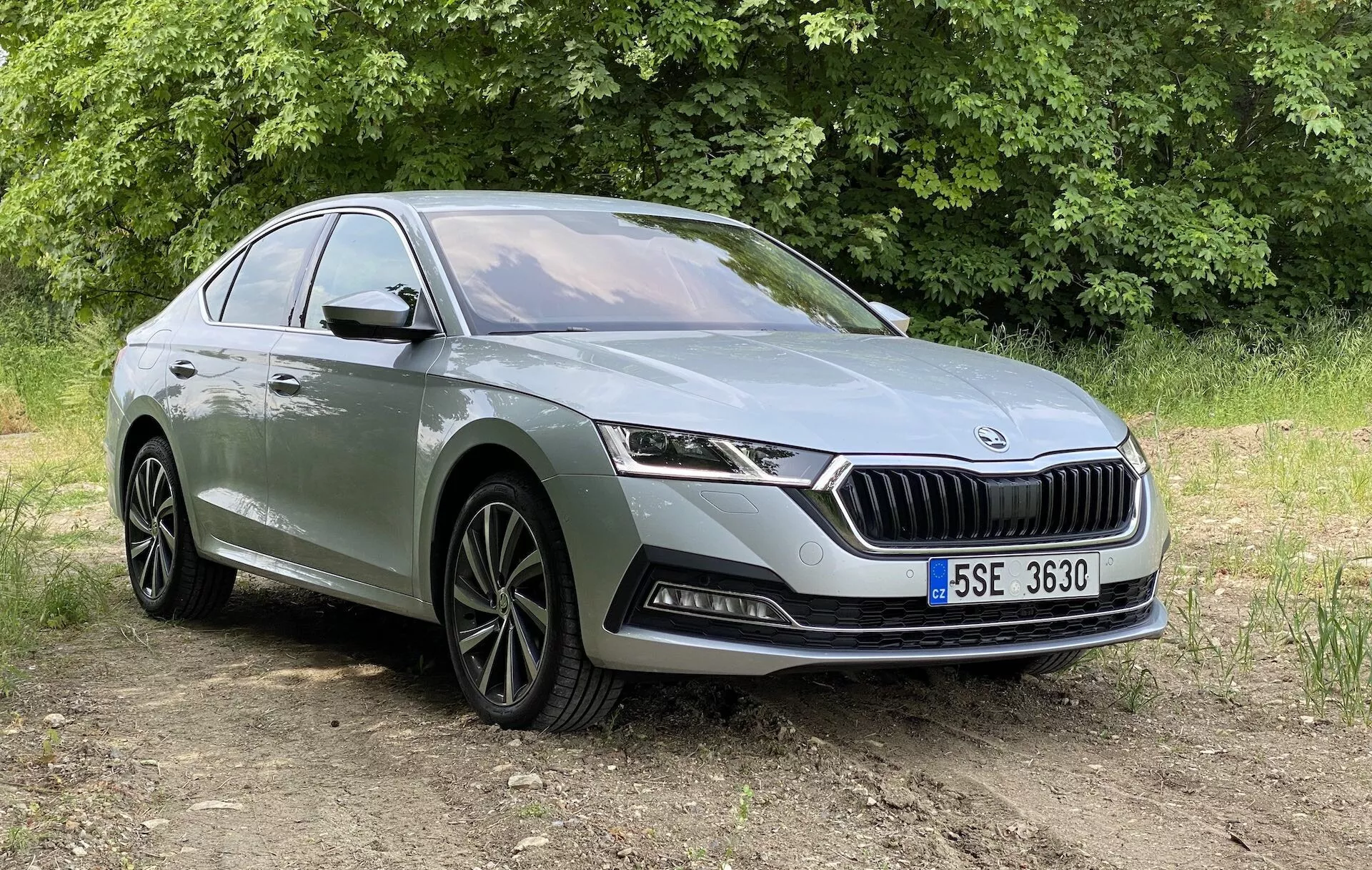 Vue avant trois-quarts d'une Skoda Octavia 2020 moderne, présentée sous un angle légèrement inférieur, mettant en avant sa façade avant, une calandre avec des lattes verticales et un logo proéminent, des phares à LED horizontaux élégants et le pare-chocs inférieur avec des phares antibrouillard et des prises d'air. La voiture est positionnée sur une surface non pavée avec de la végétation en arrière-plan, sous la lumière naturelle du jour. Les autres détails visibles incluent des jantes en alliage avec un design à multiples rayons, une ligne de caractère courant le long du profil latéral et des poignées de porte. Convient pour présenter le design automobile et les fonctionnalités des véhicules modernes dans un environnement extérieur.