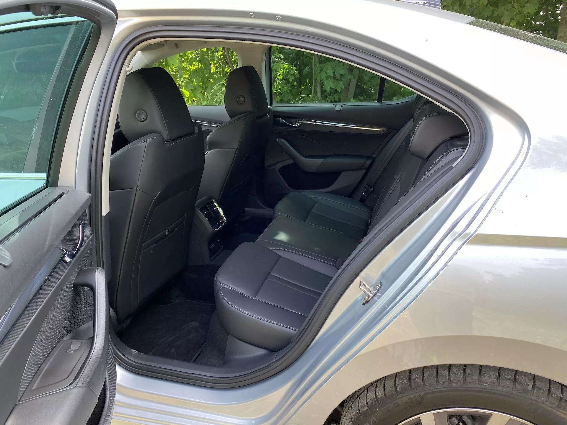 Image de l'intérieur de la Skoda Octavia 2020, axée sur le compartiment arrière des passagers. La porte arrière est ouverte, dévoilant des sièges en cuir noir, des appuie-têtes, des ceintures de sécurité et un accoudoir central replié. Le panneau de porte est fabriqué avec un matériau semblable au cuir pour plus de confort. Les éléments visibles comprennent des poches de rangement au dos des sièges avant, un tapis propre et un point d'ancrage pour siège enfant. L'éclairage ambiant suggère une journée ensoleillée dans une zone résidentielle verdoyante.