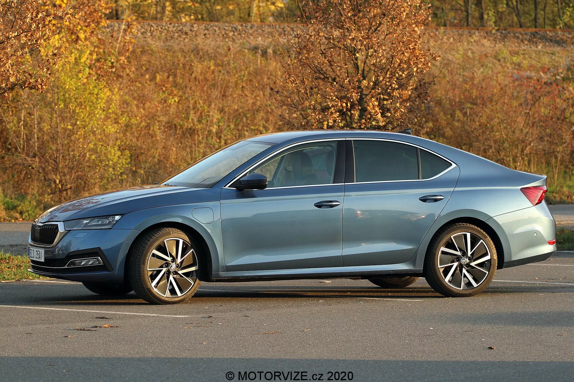Skoda Octavia 2020 berline moderne garée en extérieur, vue latérale trois quarts montrant le profil complet, la longueur et la hauteur; détails visibles comprenant des phares aigus, une ligne de toit inclinée, des lignes de caractère, des fenêtres avec des garnitures chromées, des roues en alliage multi-rayons, un environnement de feuillage automnal et une illumination naturelle de jour.