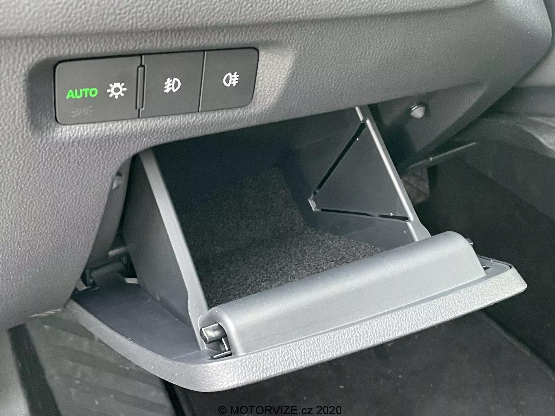 Gros plan de l'intérieur de la Skoda Octavia 2020, se concentrant sur la boîte à gants ouverte et les commandes du tableau de bord. L'angle frontal met en évidence la porte de la boîte à gants ouverte vers le bas et les boutons éclairés des phares, des feux de brouillard et des commandes d'éclairage situés au-dessus.