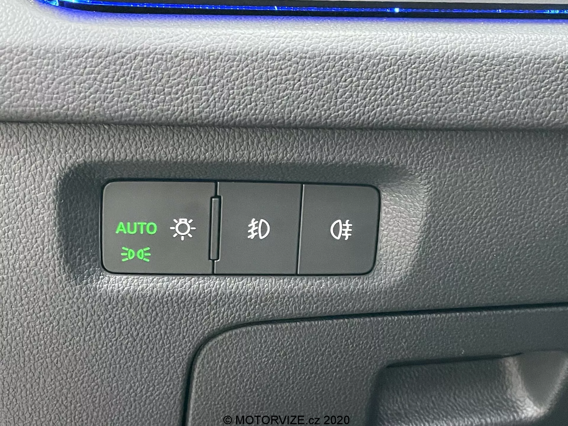 Vue rapprochée du panneau de commande de la Skoda Octavia 2020, mettant en évidence les interrupteurs des phares automatiques, des feux de stationnement et des antibrouillards. Le bouton AUTO est illuminé en vert, indiquant l'activation des phares automatiques. Le panneau en plastique texturé présente un design bicolore avec un possible reflet bleu en haut, dans un agencement intérieur de voiture.