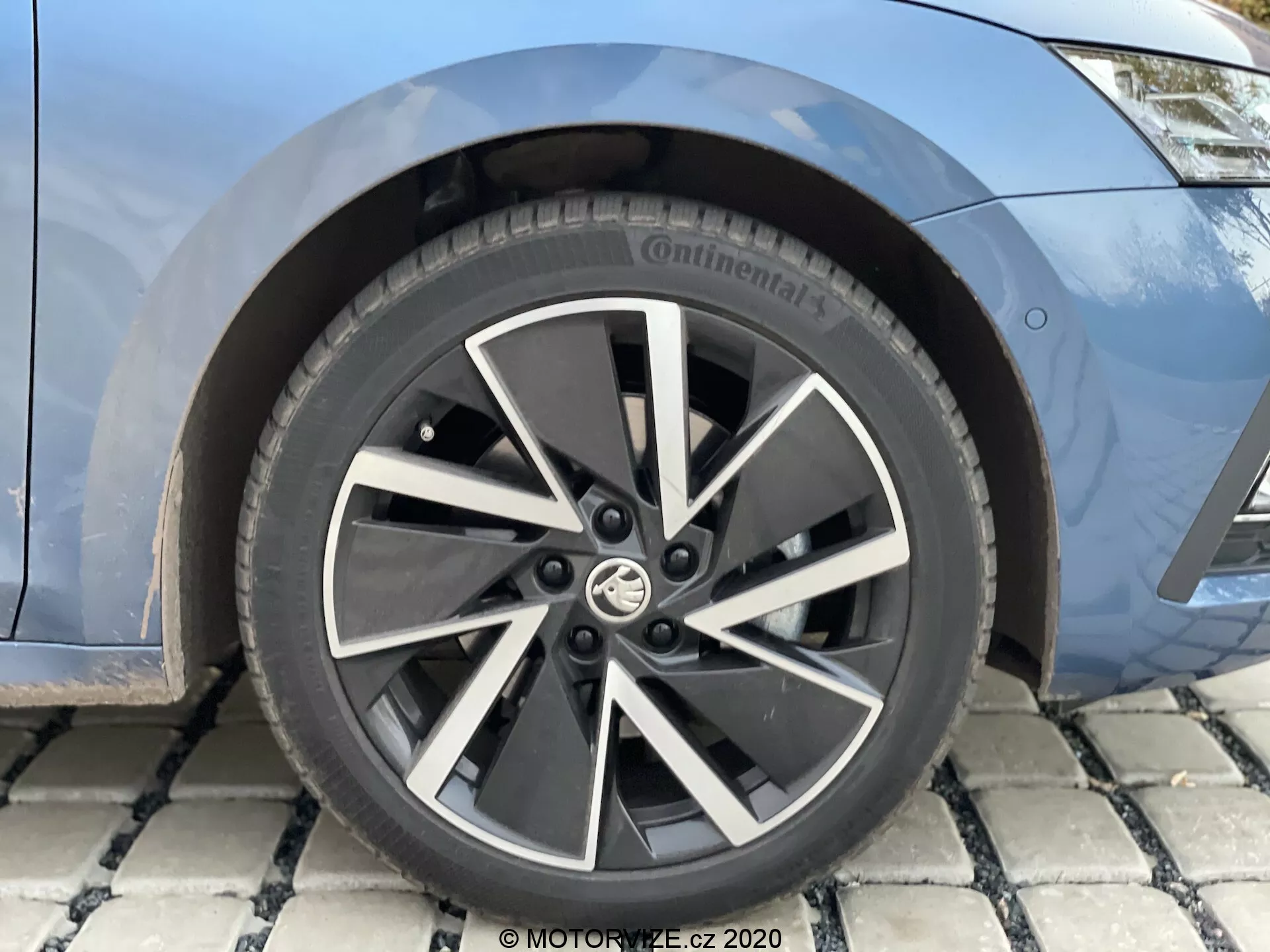 Gros plan sur la roue avant et une vue partielle de l'aile avant d'une Skoda Octavia 2020 limousine. La roue présente un design moderne à plusieurs branches en alliage bicolore noir et argent, avec l'insigne du fabricant au centre. Le pneu est de la marque Continental, probablement de la série Conti, avec des spécifications de taille alphanumériques partiellement visibles. Derrière les branches, on aperçoit le disque de frein et éventuellement l'étrier de frein, indiquant un système de freinage moderne. La carrosserie est visible en peinture bleue métallisée.