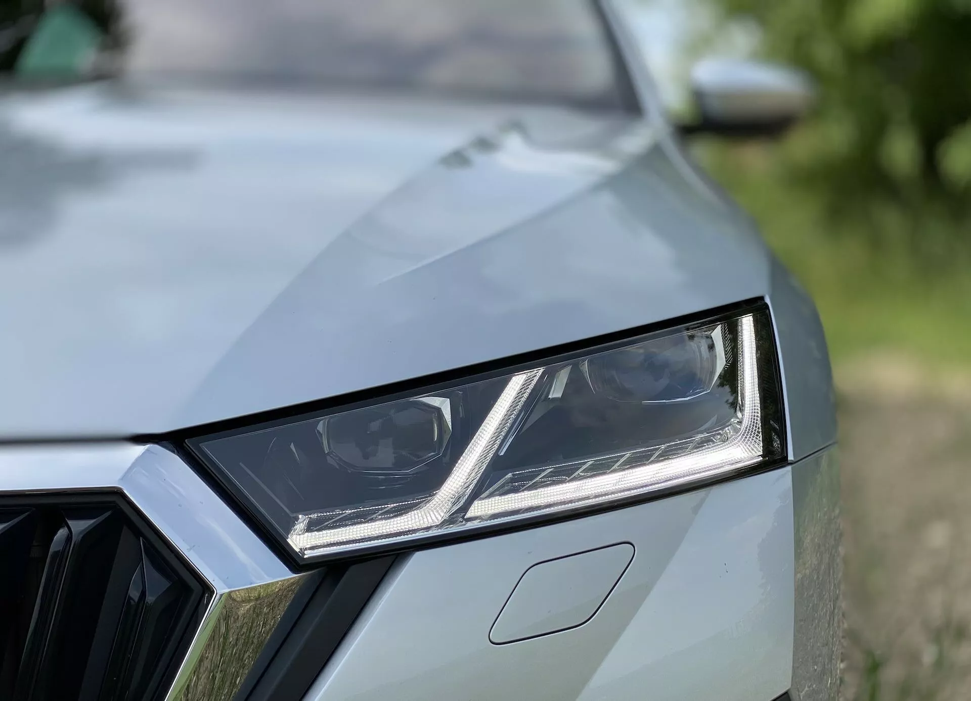 Vue rapprochée du côté avant gauche d'une voiture moderne montrant des éléments de design détaillés tels que le phare LED angulaire avec feux de jour intégrés, une vue partielle de la grille à motif géométrique et le capot avec des plis distinctifs. La finition de peinture de haute qualité et l'arrière-plan flou accentuent l'esthétique sportive et contemporaine de la voiture.