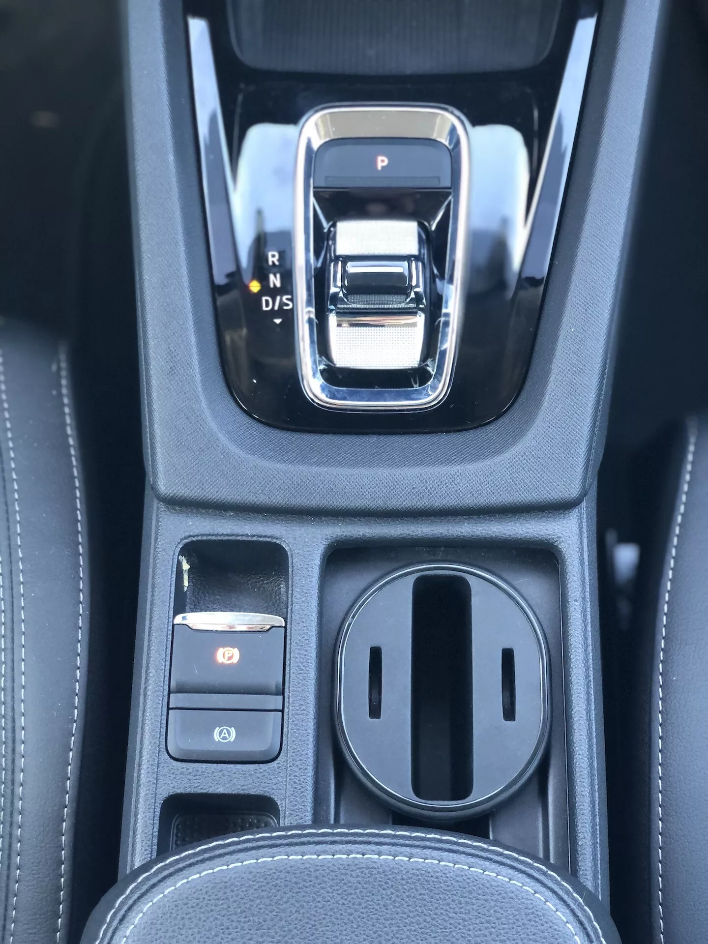 Gros plan sur la zone du levier de vitesses automatique dans une Škoda Octavia Limousine 2020. Le levier de vitesses est une manette électronique argentée et noire avec les options P, R, N et D/S. À gauche, deux boutons de commande sont visibles, avec le bouton supérieur éclairé en rouge, probablement le frein de stationnement électronique. À droite se trouve un compartiment de rangement couvert avec deux porte-gobelets. La console noire présente une texture similicuir avec des coutures détaillées, indiquant un intérieur haut de gamme avec des systèmes de transmission avancés et des systèmes de stationnement électroniques.