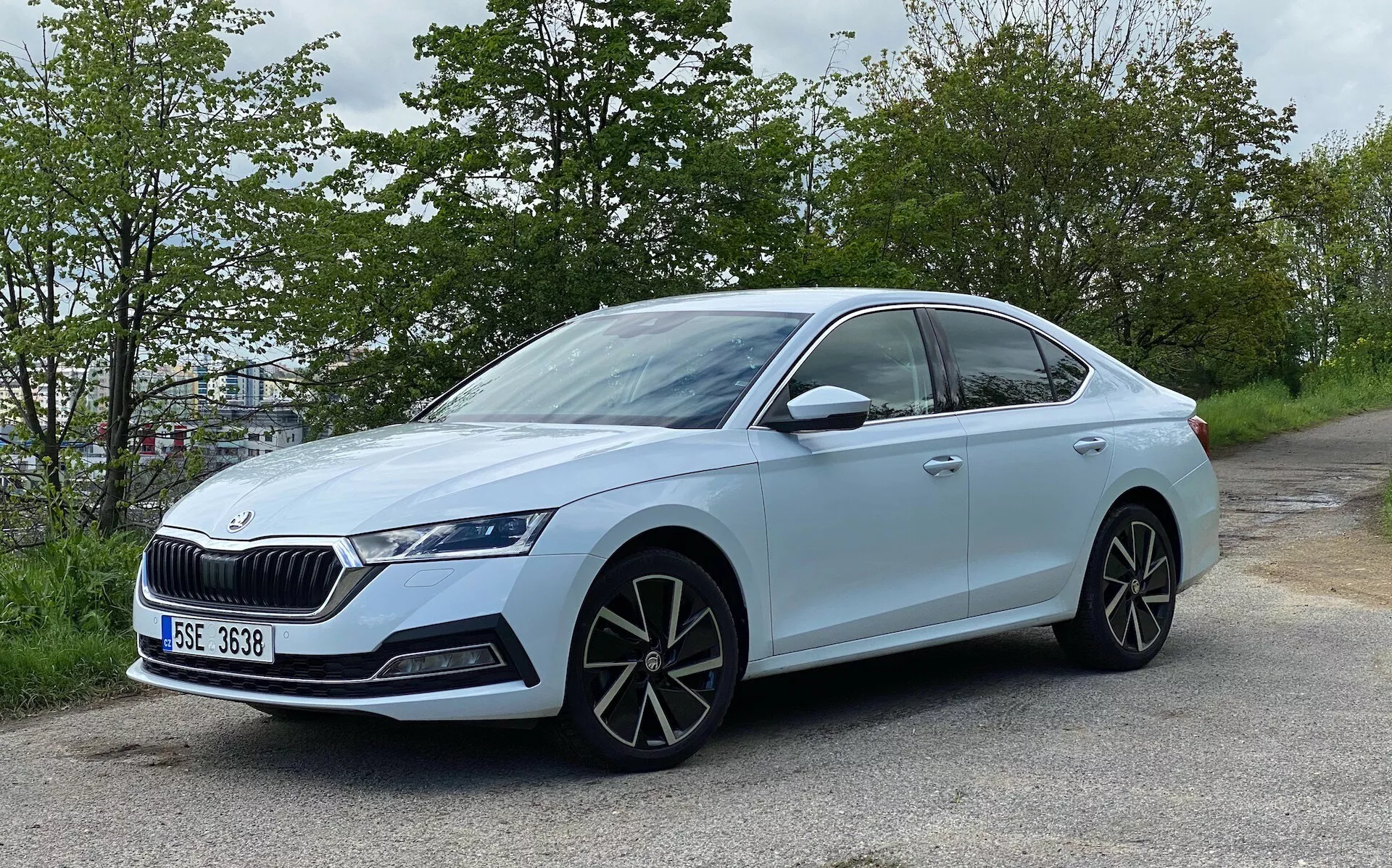 Vue latérale modérément angulée d'une berline Skoda Octavia 2020 bleu clair, positionnée au bord de la route avec un feuillage vert en arrière-plan. Montre un design moderne avec une carrosserie à quatre portes, une calandre proéminente, des phares LED agressifs, de grandes roues en alliage à rayons multiples, des rétroviseurs latéraux de la couleur de la carrosserie avec des clignotants intégrés, une ligne de toit en pente, des fenêtres teintées avec une garniture chromée, des poignées de porte affleurantes, des pneus à profil bas et un badge indiquant le modèle ou le paquet technologique.