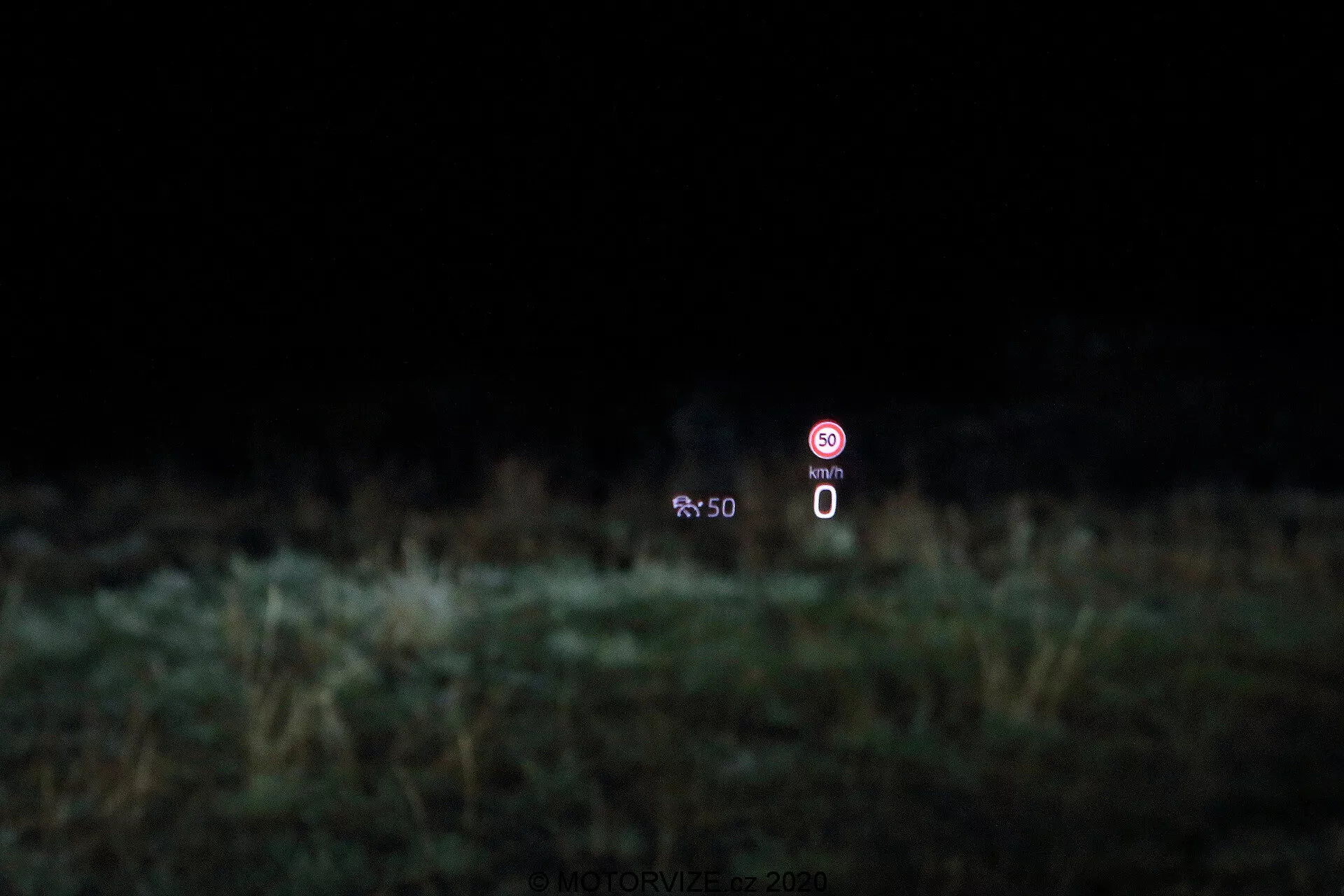 Image montrant la technologie head-up display (HUD) dans une Škoda Octavia 2020 limousine. L'affichage est projeté sur le pare-brise, montrant un compteur de vitesse statique à 0 km/h et une icône de prohibition indiquant une fonction désactivée, sur un fond flou de végétation.