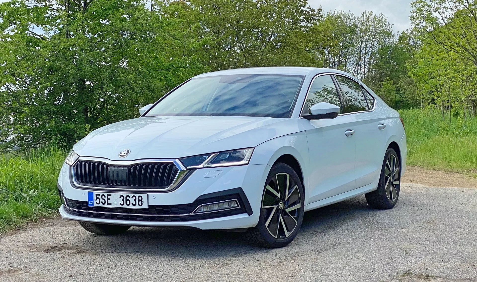 Image d'une Škoda Octavia 2020 gris bleu clair positionnée sur une route pavée avec du feuillage en arrière-plan. Prise sous un angle de trois quarts avant, la photo met en valeur les détails de conception : la calandre avant proéminente avec des lamelles verticales et l'emblème Škoda, les phares LED angulaires élégants avec feux diurnes, les feux antibrouillard séparés, les grandes jantes en alliage multi-branches, les rétroviseurs latéraux avec clignotants intégrés, les éventuels capteurs de stationnement avant et les lignes dynamiques du capot et des côtés. La scène suggère un environnement diurne serein avec un éclairage naturel.