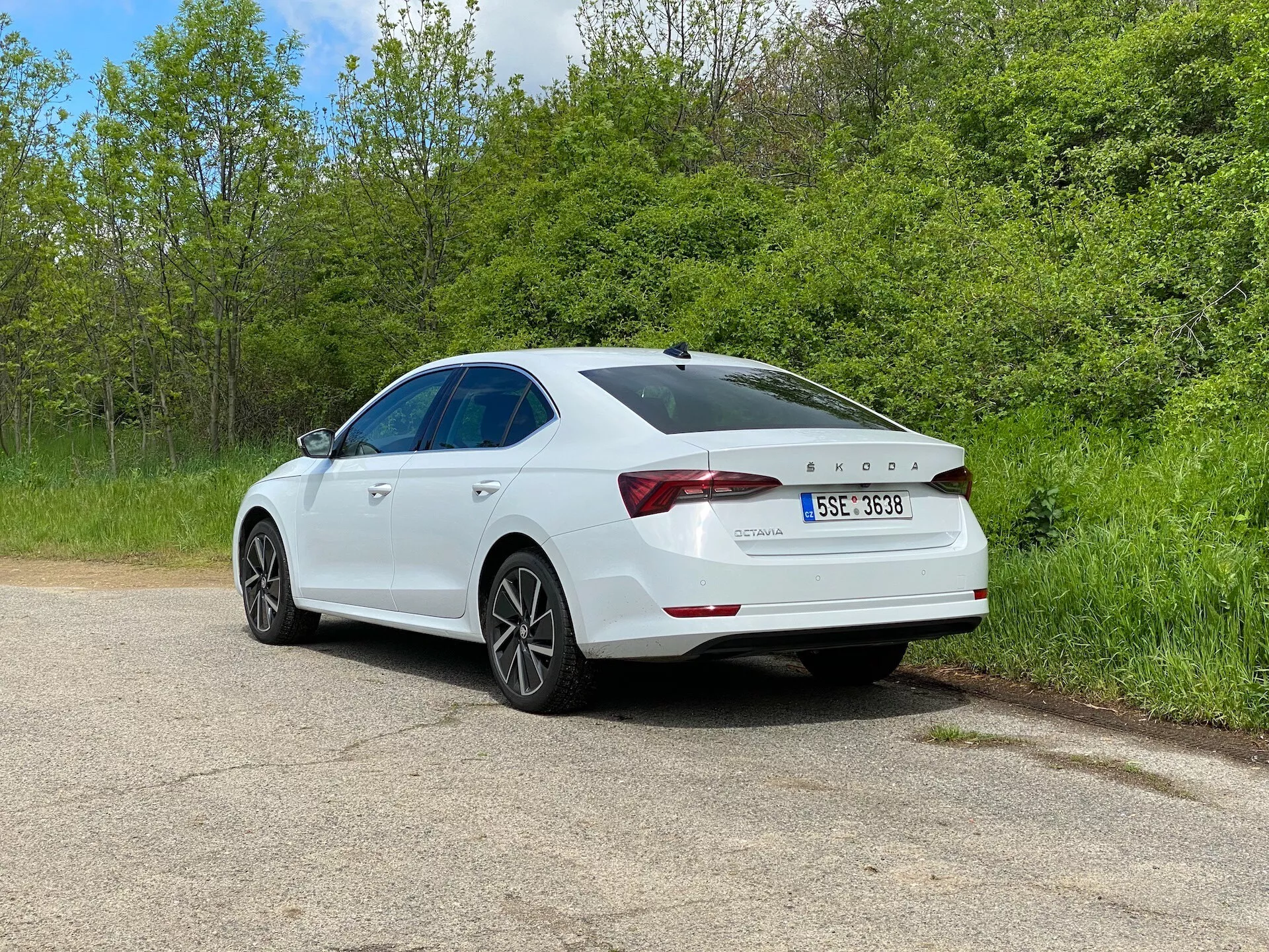 Berline blanche Skoda Octavia 2020 en vue arrière trois quarts, stationnée sur une route de gravier, entourée de verdure, avec vue détaillée sur les feux arrière distinctifs, le becquet de coffre arrière, les jantes en alliage et un toit ouvrant visible, soulignant son design moderne et ses caractéristiques technologiques, y compris le capteur de stationnement sur le pare-chocs.