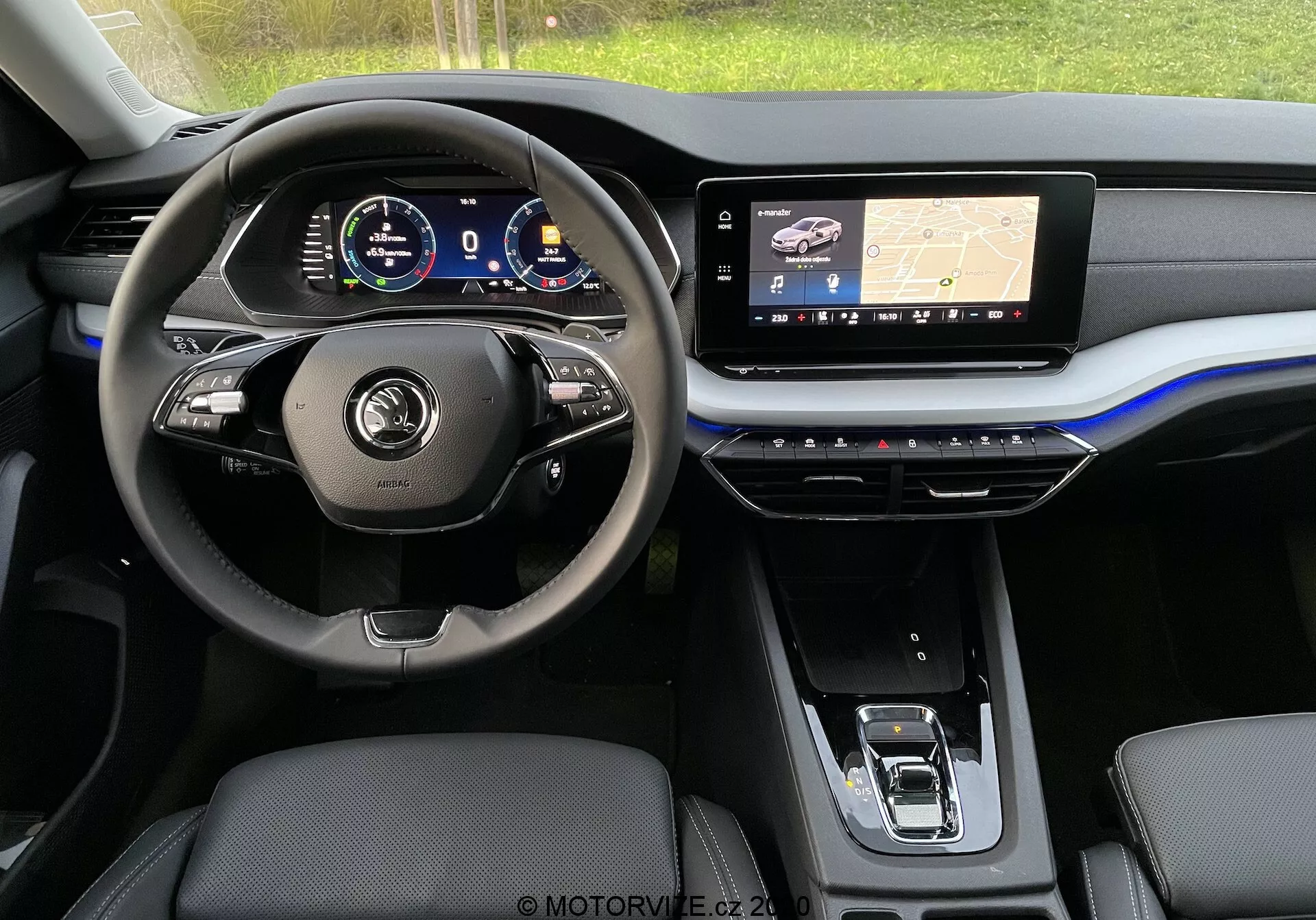 Vue intérieure d'une Skoda Octavia Limousine 2020 moderne depuis la banquette arrière, montrant le tableau de bord avant et se concentrant sur la zone du siège conducteur. Le volant est équipé de boutons multifonctions et d'un revêtement en cuir, derrière lequel un tableau de bord numérique affiche divers indicateurs du véhicule. Le tableau de bord central comprend un grand écran tactile d'infodivertissement affichant une carte de navigation avec technologie GPS, tandis que la console centrale en dessous contient des boutons tactiles pour la régulation du climat. Sont visibles les sièges avant garnis de cuir ou similaires et un sélecteur de vitesse électronique shift-by-wire. L'esthétique est élégante avec des tons sombres, accents métalliques et un audacieux soulignement bleu sur le tableau de bord.