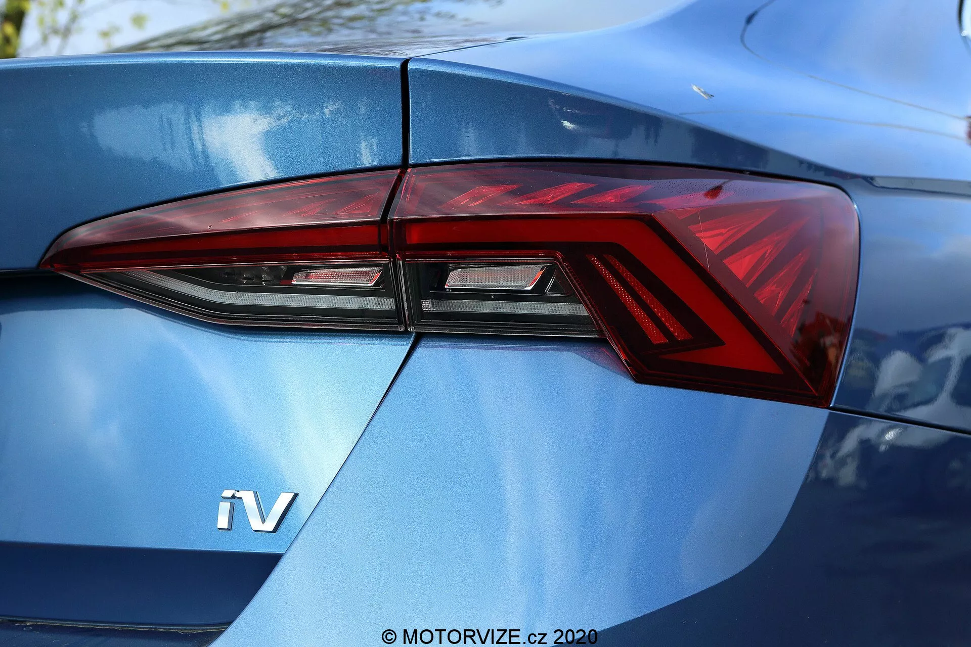 Détail en gros plan de la section arrière d'une Škoda Octavia Limousine 2020, focalisé principalement sur l'ensemble du feu arrière gauche vu sous un angle de trois quarts. Le design moderne du feu arrière présente des angles vifs et des éléments LED distinctifs, intégrés de manière transparente aux lignes de carrosserie de la voiture. Un badge 'iV' visible sur le coffre bleu métallisé brillant suggère un véhicule hybride ou électrique.