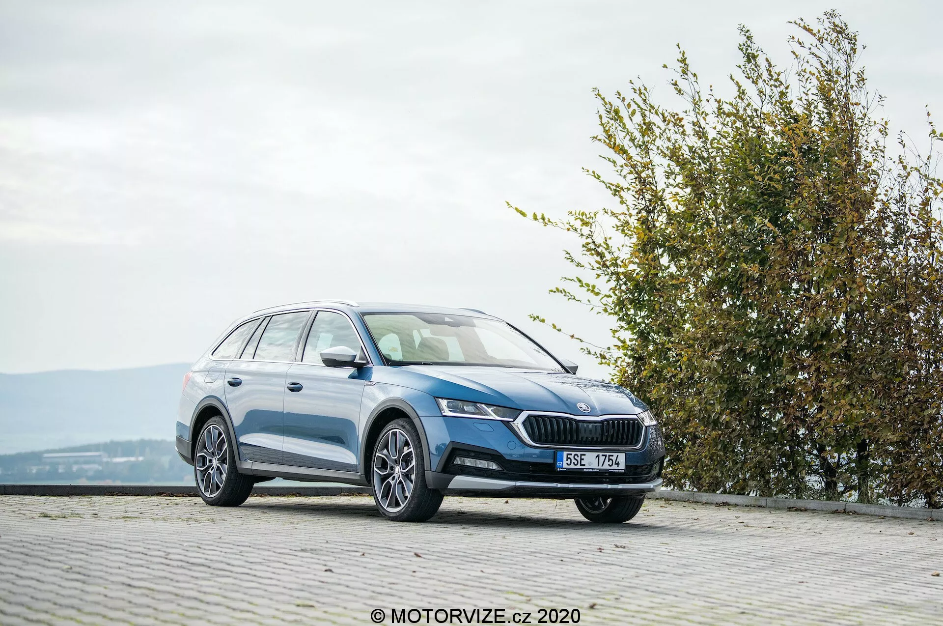Grand break 2020 Skoda Octavia Scout, photographié en vue de trois quarts avant sur une zone pavée de style pavé à un endroit élevé. L'image montre la calandre avant avec un emblème proéminent, des phares angulaires élégants, des feux antibrouillard intégrés, un profil latéral lisse avec des roues en alliage, des rails de toit et une finition peinture brillante. Le cadre environnant comprend une haie ou un buisson à droite, suggérant la proximité d'un parc ou d'un espace vert, dans des conditions météorologiques favorables.