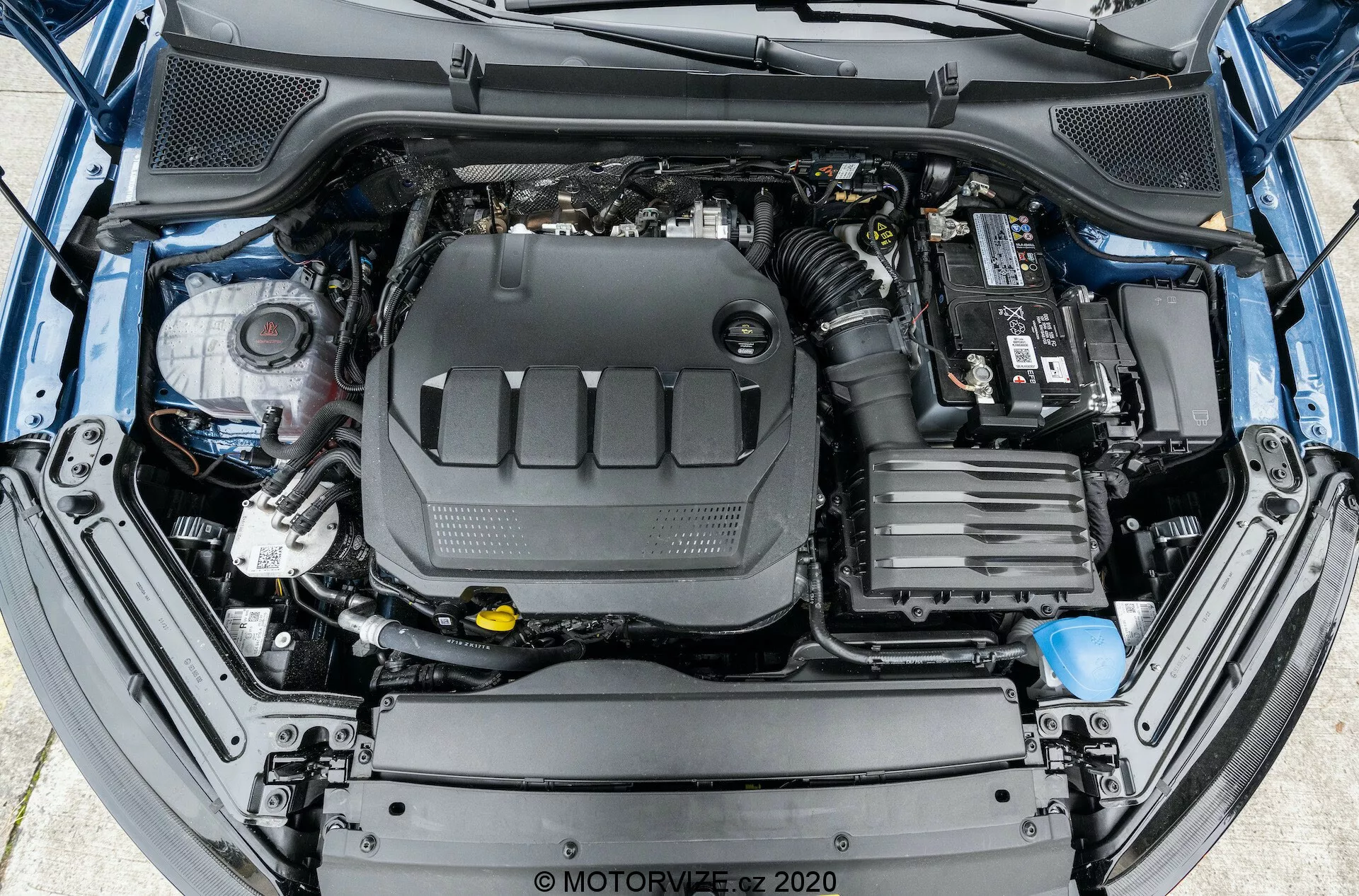 Vue plongeante sur le compartiment moteur ouvert d'une voiture, montrant une disposition complète du moteur et de ses composants environnants. Le bloc moteur est positionné au centre, couvert par un capot en plastique. À droite, la batterie de la voiture est visible avec les connexions des bornes sur le dessus. Vers l'avant, les composants du système d'admission d'air du véhicule sont visibles. Sur le côté gauche, divers réservoirs de fluides, comme le réservoir de liquide de frein avec un couvercle jaune, sont présents. Des tuyaux et des faisceaux de câbles sont également visibles dans tout le compartiment moteur. La structure du pare-chocs avant et de la grille est partiellement visible sur le bord inférieur de l'image.