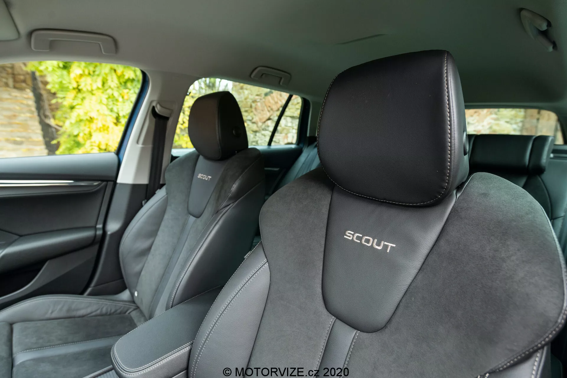 Vue intérieure d'une Skoda Octavia Scout 2020 depuis la banquette arrière, montrant les sièges avant. L'angle favorise le côté gauche, offrant une vue dégagée sur le siège du conducteur avec un logo embossé 'SCOUT'. Les sièges sont garnis d'un design bicolore, principalement noir avec des accents gris clair et un matériau central type suédine, soulignant un design sportif. L'image montre aussi le montant B, des garnitures intérieures sombres, un fragment de porte avant, une grille de haut-parleur et une poignée de porte chromée. L'arrière-plan extérieur visible à travers les fenêtres semble montrer du feuillage, indiquant que la voiture est stationnée en extérieur avec une lumière naturelle.