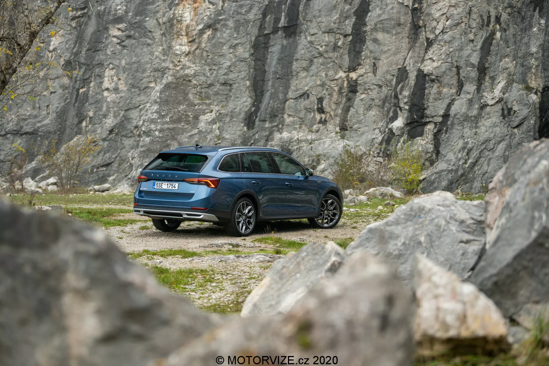 L'image montre une voiture Skoda Octavia Scout 2020 garée sur un terrain rocheux, avec une carrière ou une falaise en arrière-plan, capturée d'une légère perspective téléobjectif. L'angle arrière trois quarts offre une vue détaillée du profil latéral et de l'arrière du véhicule, montrant le design moderne du break, la ligne de toit arrière inclinée et la peinture bleu foncé. Les éléments visibles incluent l'assemblage des feux arrière, la forme des fenêtres, l'essuie-glace arrière, la conception des roues et les rétroviseurs latéraux. L'environnement évoque des thèmes d'aventure et d'exploration, indiquant que la voiture est apte à atteindre des endroits reculés et des terrains accidentés.