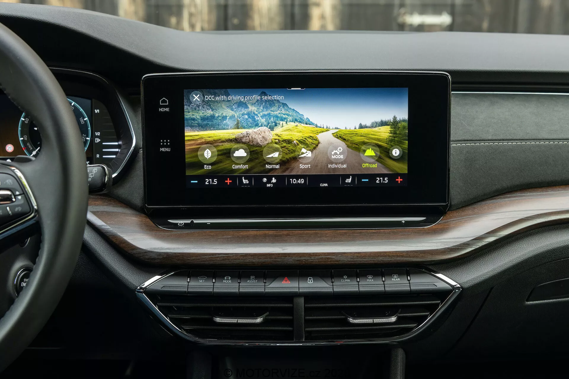 Vue intérieure de la Skoda Octavia 2020 (Octavia Scout) mettant en avant le système d'infodivertissement moderne et le tableau de bord, photographiée depuis la perspective du compartiment passagers arrière. L'écran tactile large et horizontal sur le tableau de bord affiche des graphiques colorés, les réglages de la climatisation, l'heure et la température extérieure. En dessous, des boutons et des cadrans de contrôle du climat sont bien rangés. Le tableau de bord luxueux présente des matériaux doux au toucher et une plaquette en bois, avec des bouches d'aération discrètement intégrées.