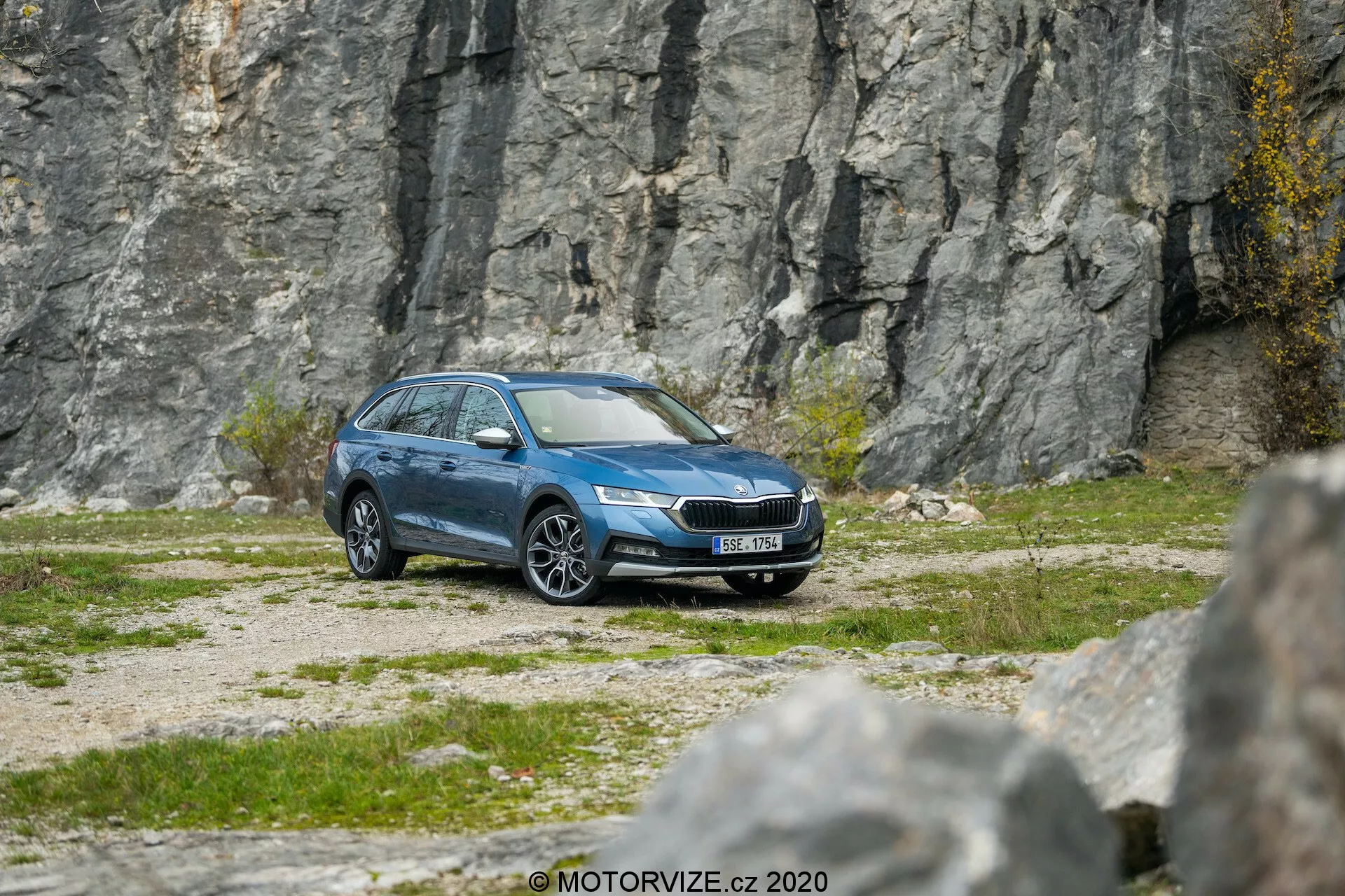 Vue avant latérale d'une break Skoda Octavia Scout 2020 bleue sur un terrain de gravier avec un fond rocheux, montrant des phares LED modernes, une calandre avant proéminente, des jantes en alliage, des rétroviseurs latéraux, des garnitures de fenêtre, des rails de toit et une finition métallique propre, suggérant des caractéristiques avancées comme la technologie d’éclairage, la surveillance de la pression des pneus et de possibles systèmes de sécurité.