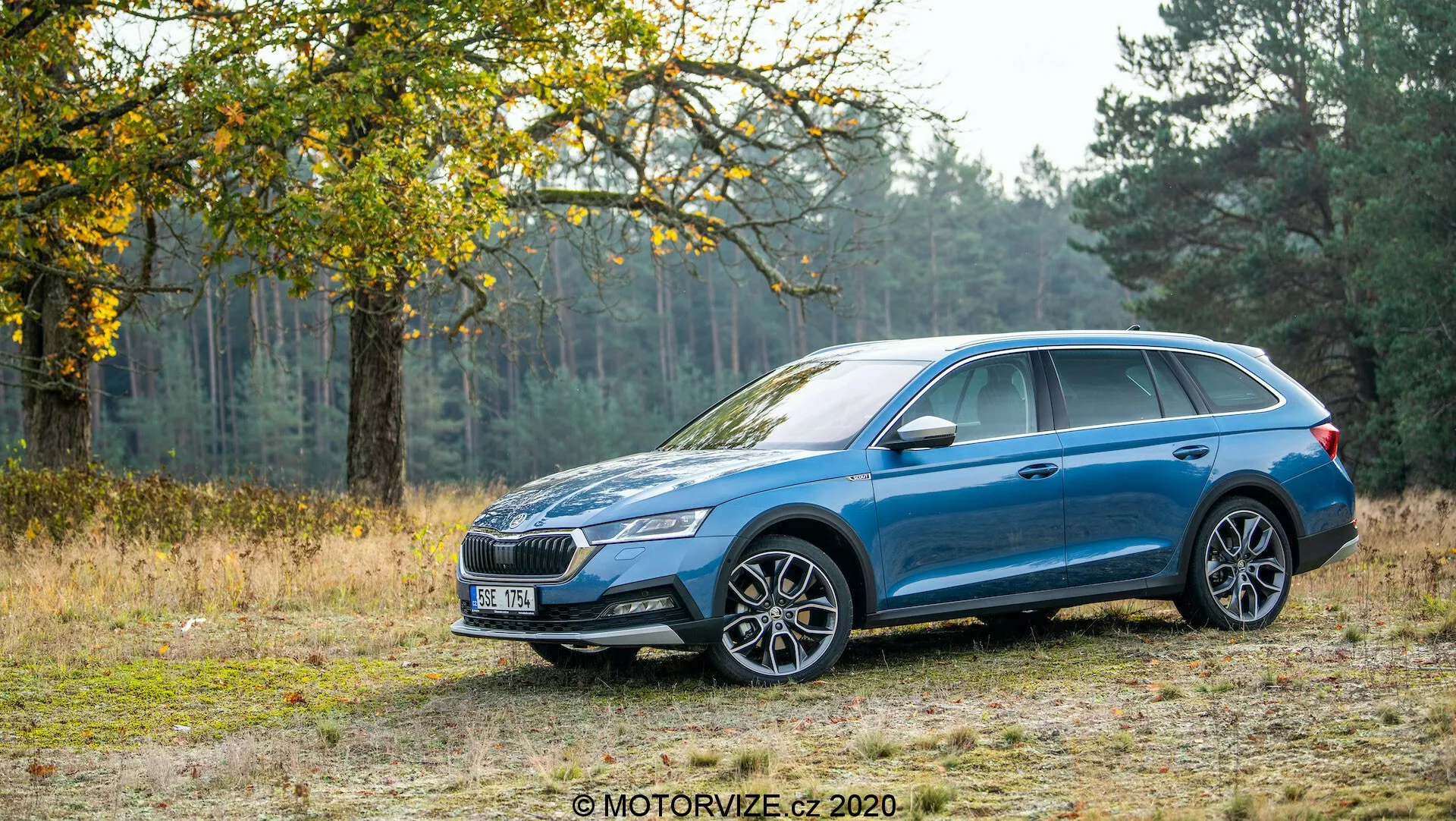 Profil latéral de la voiture familiale bleue Skoda Octavia Scout 2020 sur une piste de terre avec arrière-plan d'arbres et d'herbe, montrant sa capacité à gérer des terrains accidentés. La voiture est dotée d'une peinture métallique, d'un design de roues en alliage à plusieurs rayons, de phares à LED, d'une calandre avant distinctive, de barres de toit, de rétroviseurs et poignées de porte couleur carrosserie et d'un toit ouvrant. Les caractéristiques de design incluent la ligne de caractère allant de l'arche de la roue avant jusqu'aux feux arrière enveloppants.