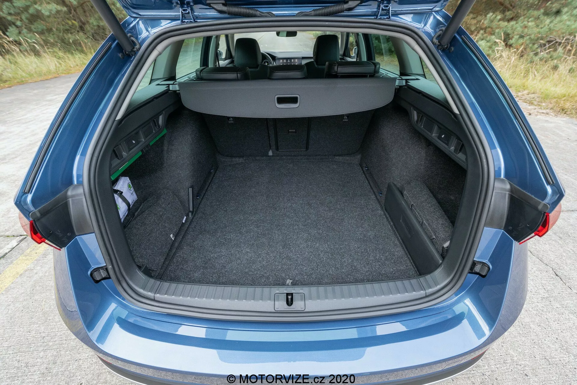 Vue rapprochée du coffre ouvert d'une moderne Skoda Octavia Scout 2020, avec un design de hayon entièrement ouvert. L'image capture le coffre du pare-chocs arrière au pare-brise arrière, y compris une partie du toit. La zone de chargement est recouverte de moquette et présente un plancher de chargement plat ainsi qu'un petit compartiment de rangement à gauche. Les sièges arrière sont en position normale avec un cache-bagages rétractable étendu, dissimulant les objets dans le coffre.
