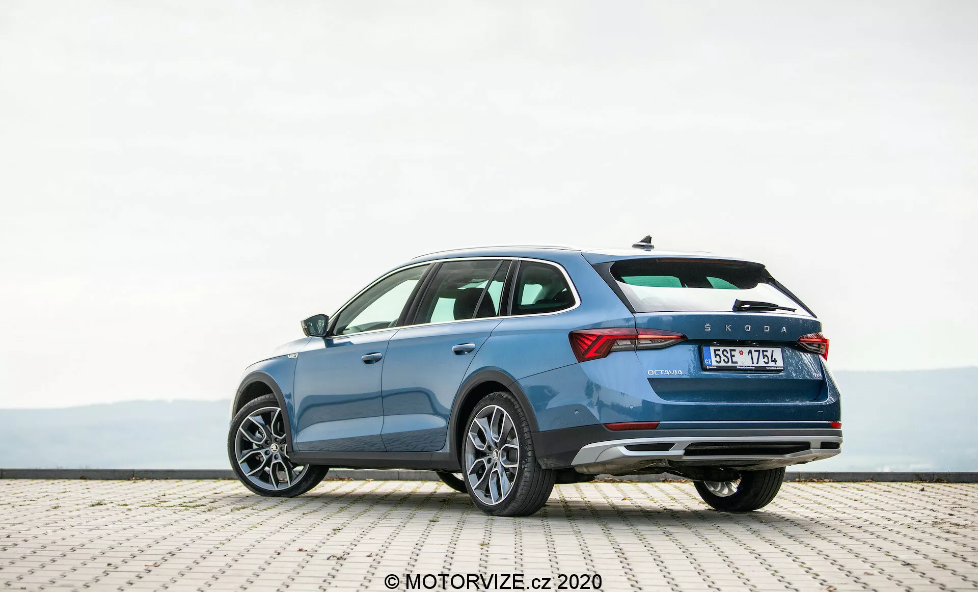Image détaillée d'une Skoda Octavia Scout 2020 bleue, garée sur une surface carrelée, prise d'une perspective arrière trois quarts. La photographie met en valeur le profil élégant du véhicule, le design distinctif du hayon et le côté droit. Les caractéristiques montrées comprennent des feux arrière horizontaux, une ligne de caractère suggérant le mouvement, un pare-chocs arrière avec des réflecteurs possiblement équipés de capteurs de stationnement, un système d'échappement double et des jantes en alliage à plusieurs rayons. L'image évoque un habitacle spacieux, des fonctionnalités de sécurité avancées et des éléments de design contemporain adaptés aux passionnés de technologie automobile.