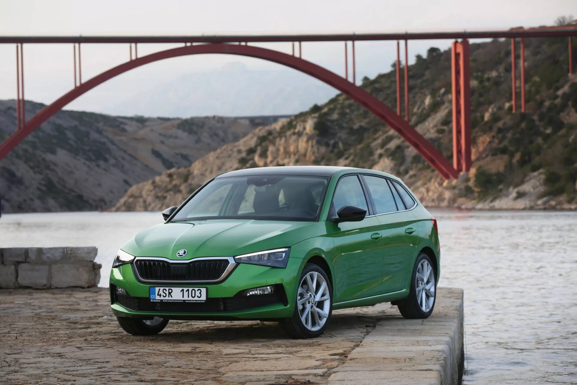 Vue avant trois quarts inclinée d'une hatchback Skoda Scala 2019 en vert, sur béton près de l'eau avec un pont rouge en arrière-plan pendant la journée, mettant en avant le design et les caractéristiques