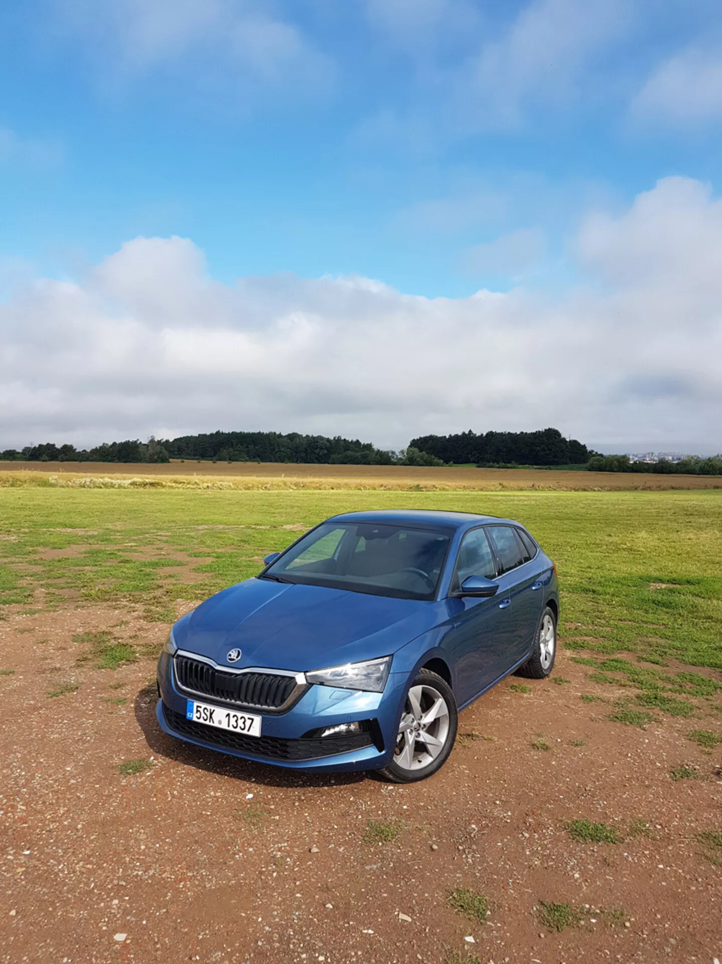 Sedan Skoda Scala 2019 bleu, garé sur l'herbe sous un ciel couvert, présentant calandre avant, phares et profil côté passager avec arrière-plan de campagne