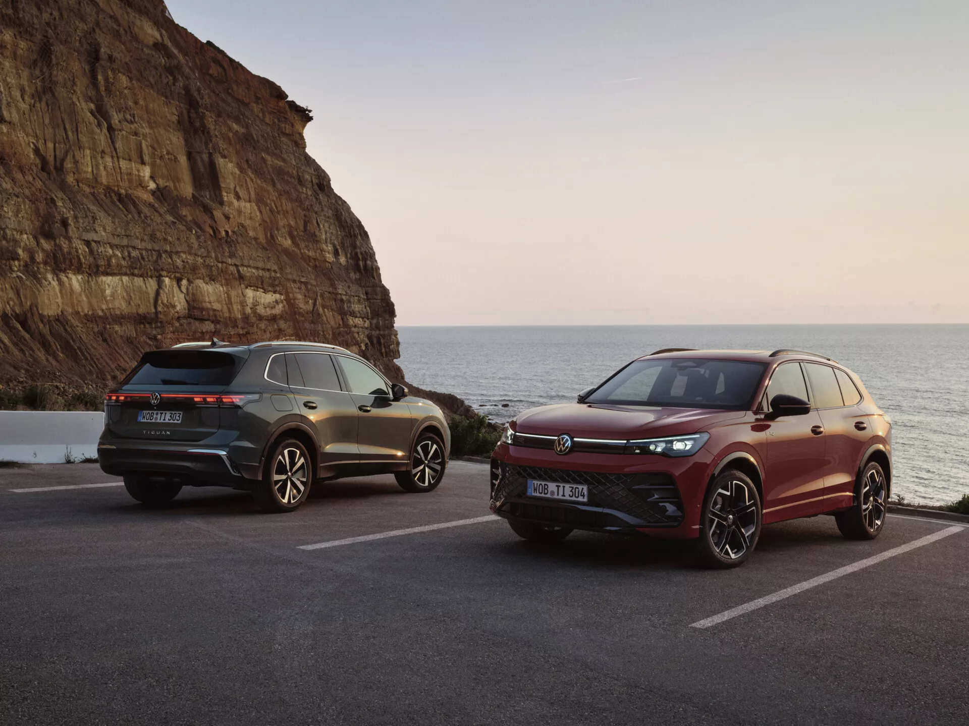 SUVs de luxe dans un cadre côtier pittoresque au coucher du soleil, mettant en vedette un modèle gris avec feux arrière LED et éléments de design sportif et un modèle bordeaux avec une grille avant orientée performance et des systèmes avancés d'assistance au conducteur