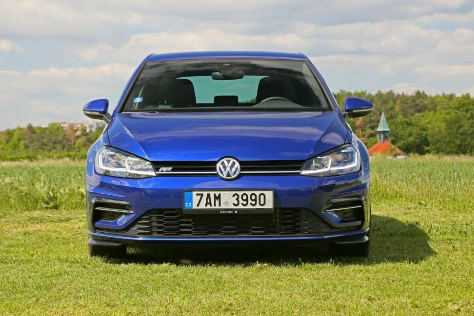 Vue de face d'une Volkswagen Golf bleue garée sur un champ herbeux sous un ciel dégagé, mettant en avant sa finition métallique, le design de sa grille, la technologie de ses phares LED et son pare-chocs avant sportif incluant les prises d'air et l'emblème.