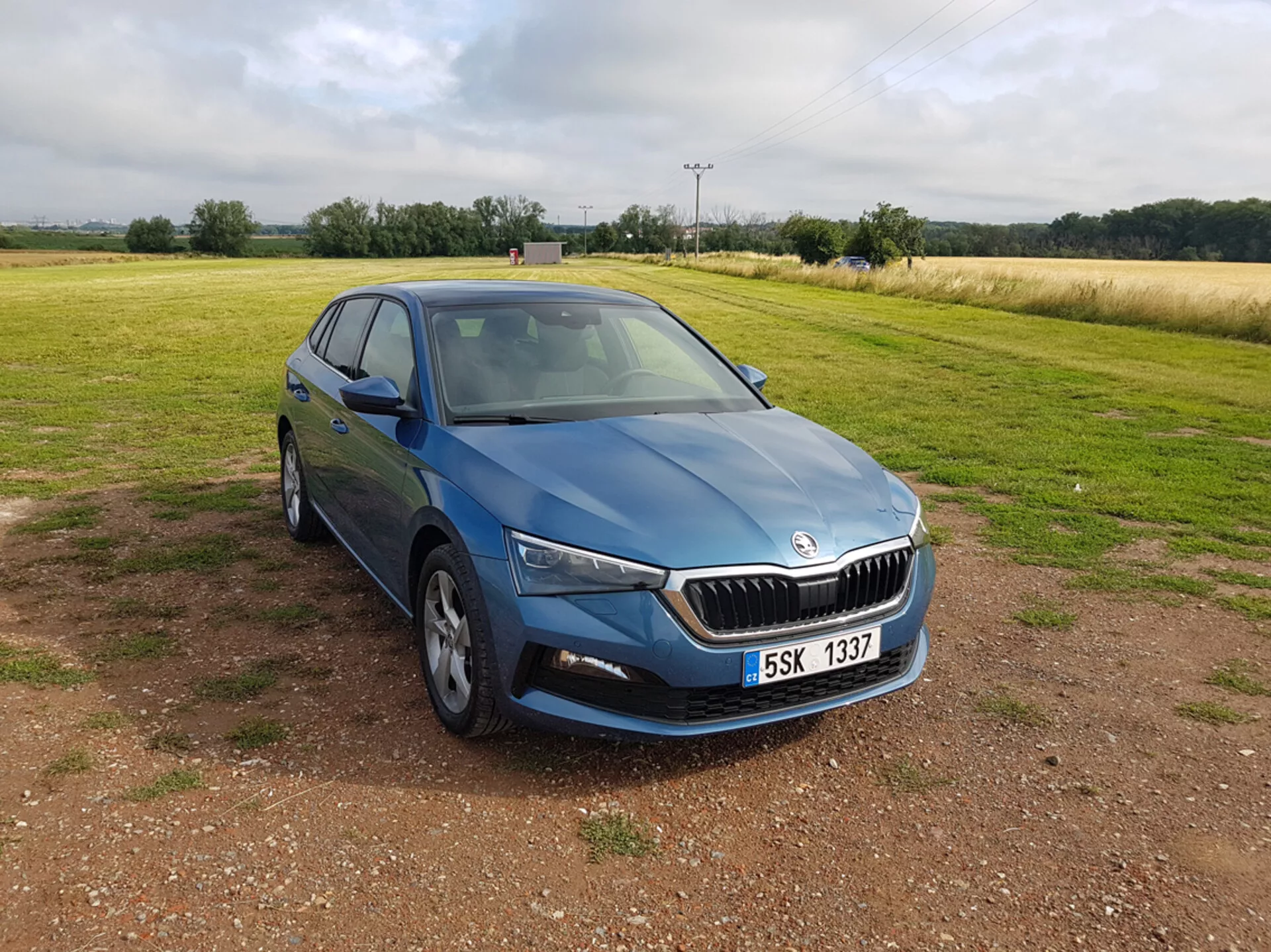 Skoda Scala bleue 2019 vue sous un angle avant trois-quarts soulignant le design, les phares LED, la grille chromée, les roues en alliage à plusieurs branches, les clignotants intégrés sur les rétroviseurs latéraux et les vitres teintées, placée sur du gravier avec un champ à l'arrière-plan sous un ciel nuageux.