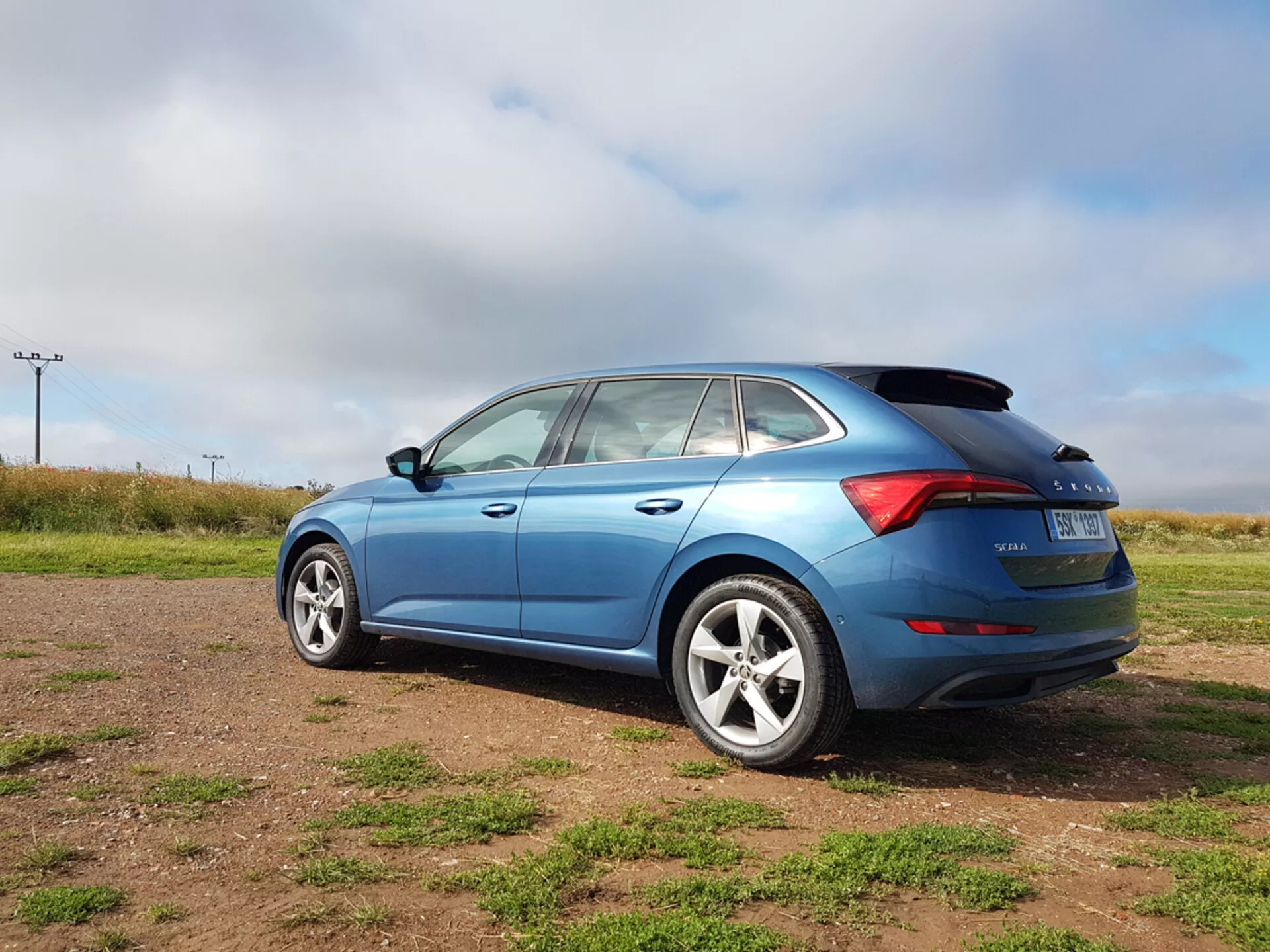 Hatchback bleu Skoda Scala 2019 montrant les éléments de design latéral et arrière, y compris la forme des fenêtres, les feux arrière LED et les roues en alliage, photographié sous un angle arrière de trois quarts sur un terrain herbeux