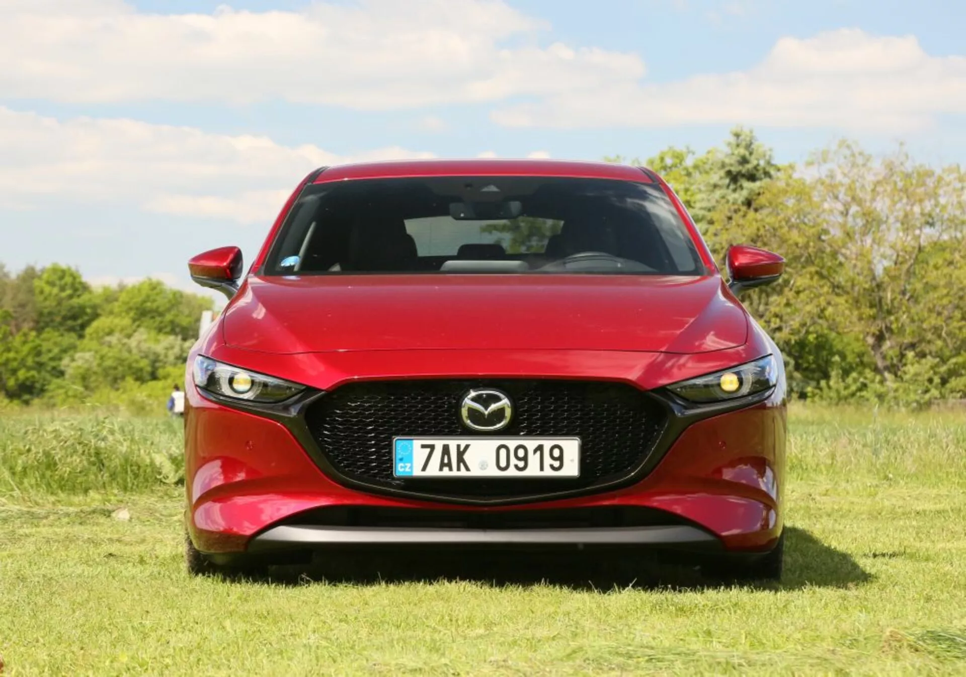 Véhicule Mazda 2019 en rouge, présentant une vue de face avec une grille proéminente comportant l'emblème de Mazda, des phares LED et un design de capot profilé. Positionné en extérieur sur l'herbe avec des arbres et un ciel bleu en arrière-plan.