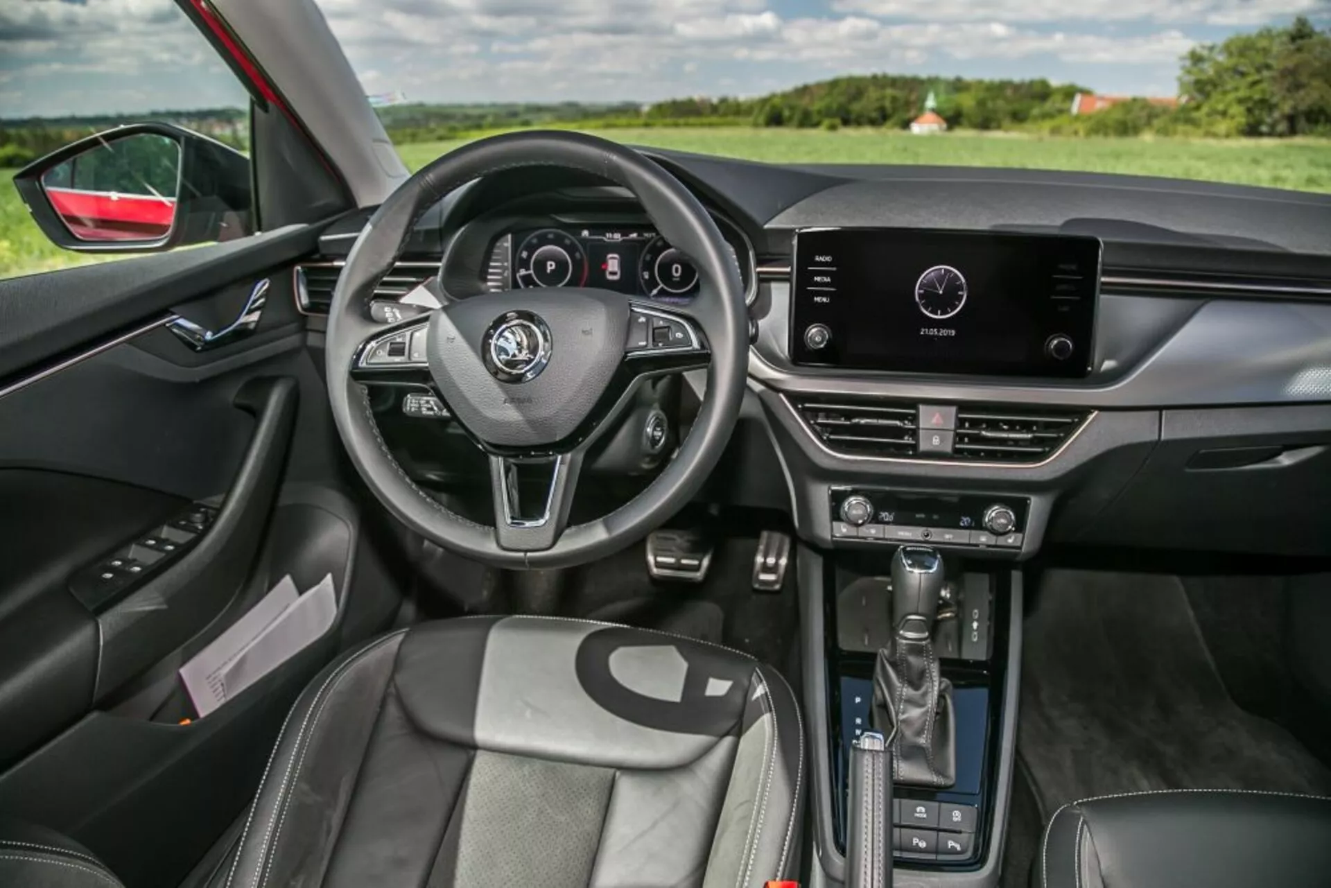 Vue intérieure de la Skoda Scala 2019 depuis le côté passager avant présentant un volant à trois branches avec commandes intégrées et tableau de bord numérique, système d'infodivertissement tactile sur la partie supérieure de la console centrale avec commandes manuelles du climatiseur en dessous, levier de vitesse automatique, sièges avant profilés avec un revêtement type cuir, panneau de porte côté conducteur avec réglages électriques des fenêtres et des miroirs, éclairage d'ambiance intérieur, rétroviseur à atténuation automatique, et une visibilité sur le paysage au travers du pare-brise et de la fenêtre côté conducteur, mettant en avant le design ergonomique et la technologie intégrée.