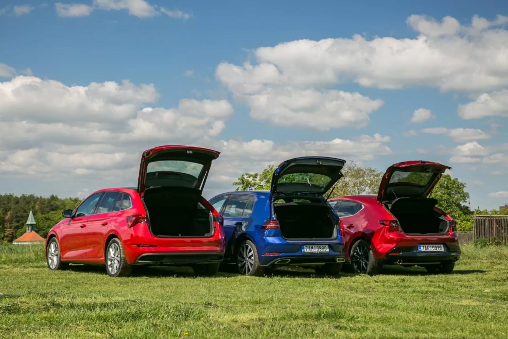 Modèles Skoda Scala 2019 en rouge et bleu garés dans un champ herbeux, présentant les profils arrière, les coffres ouverts pour la fonctionnalité de l'espace de chargement et les détails de conception sous un ciel clair.