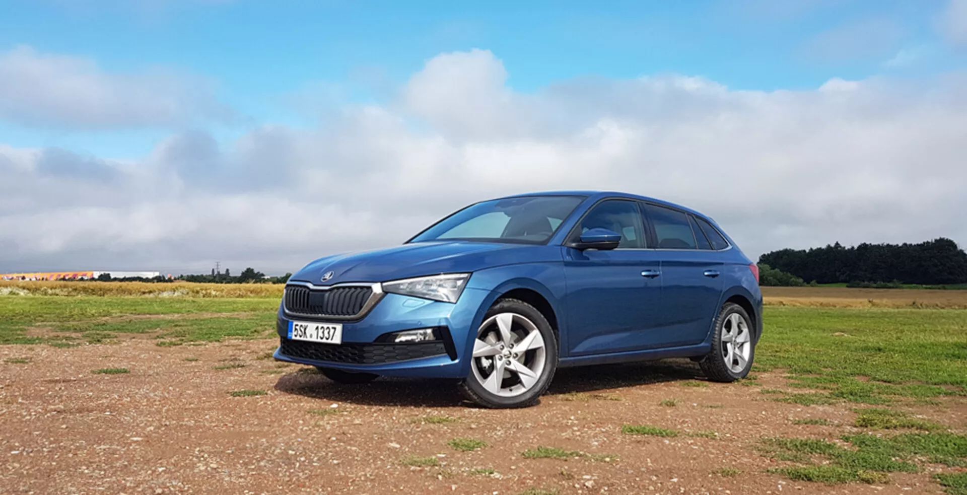 Hatchback bleu Skoda Scala 2019 sur surface non pavée avec grille avant distinctive, phares LED modernes, jantes en alliage et profil latéral gauche élégant sur fond de ciel partiellement nuageux
