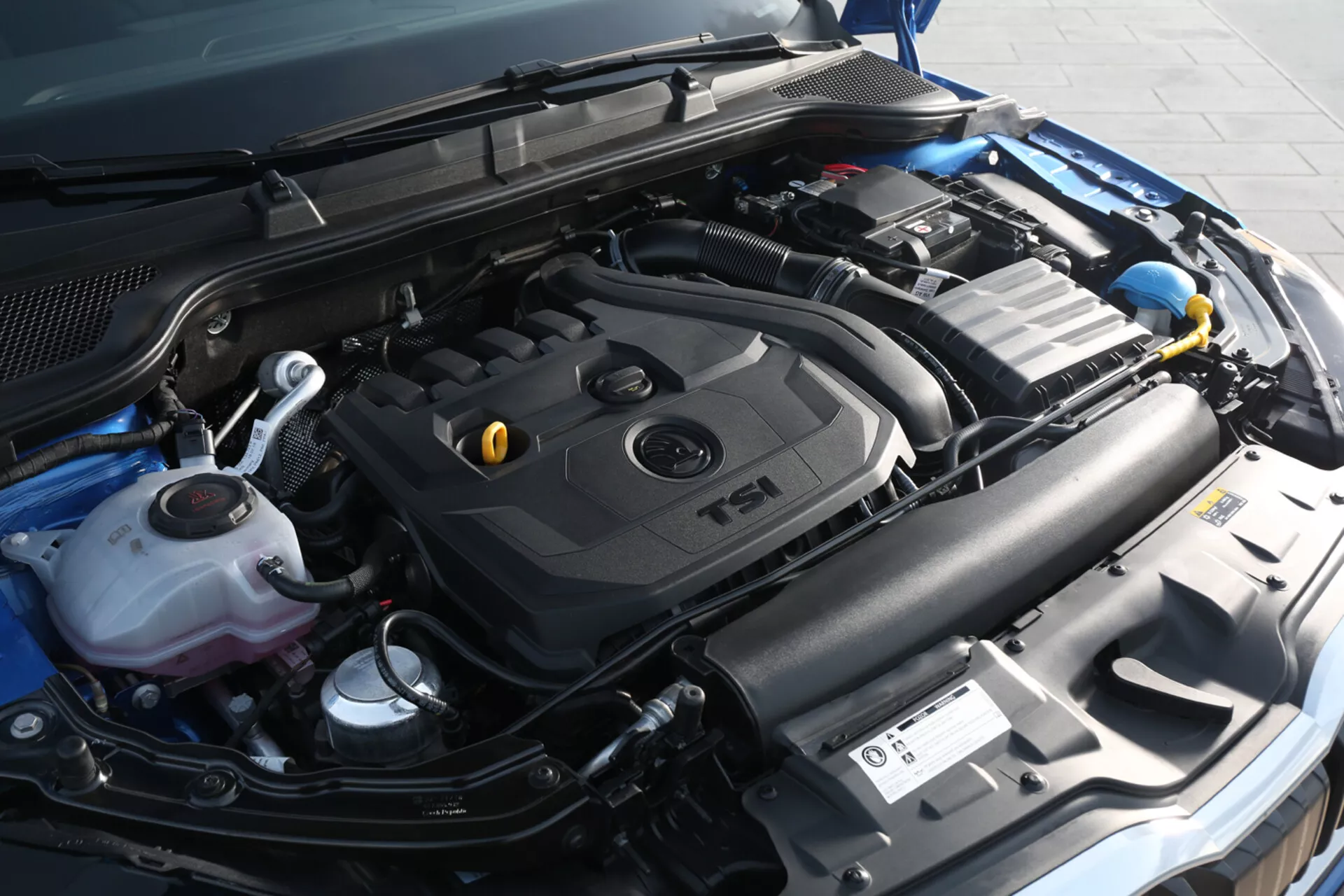 Vue rapprochée du compartiment moteur d'une Skoda Scala 2019, mettant en avant le couvercle de moteur TSI turbocompressé et les composants incluant le réservoir de liquide de refroidissement, le conteneur de liquide lave-glace, la batterie de la voiture et le système d'admission depuis un angle frontal légèrement à droite