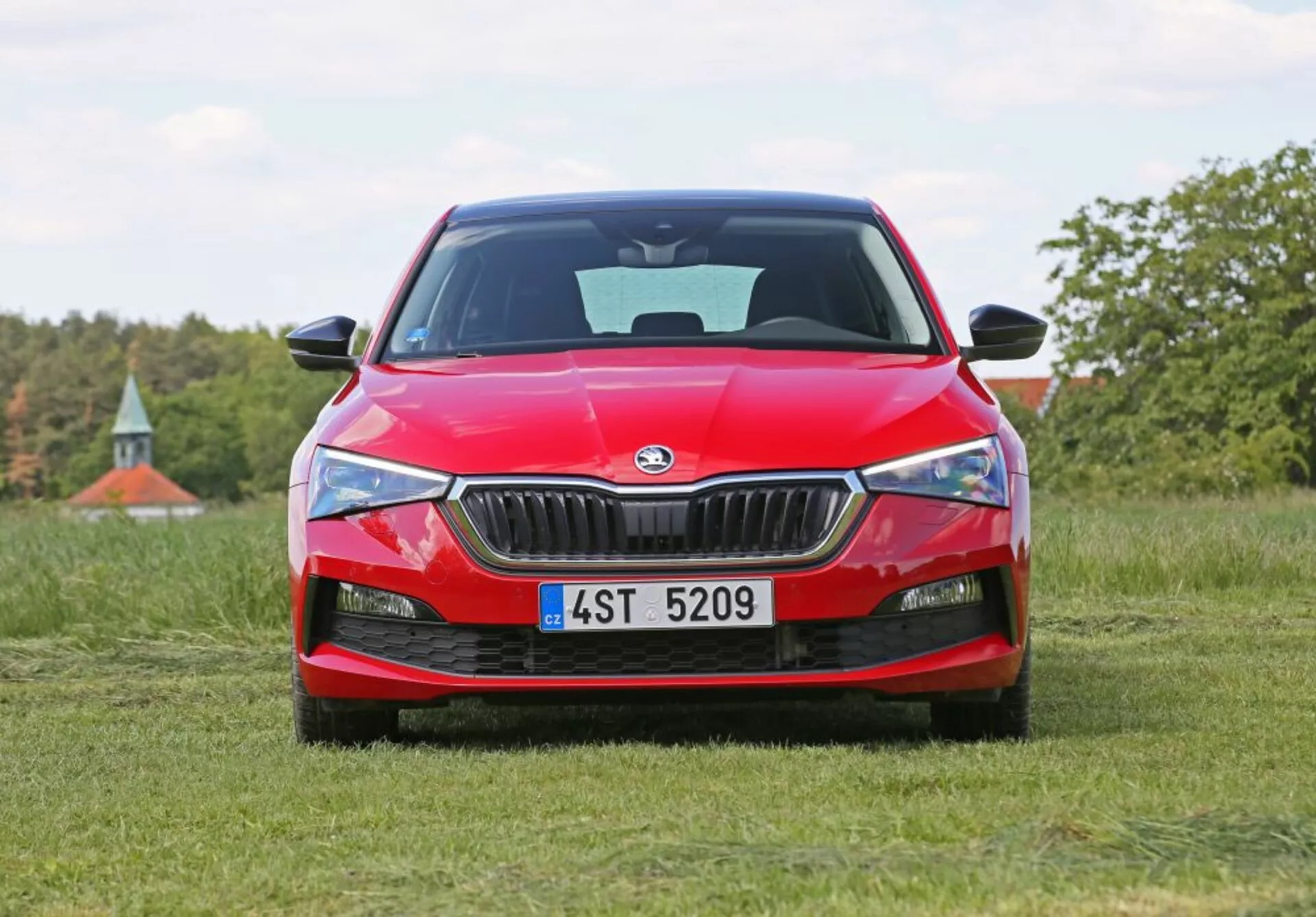 Skoda Scala 2019 en rouge positionnée sur un champ herbeux avec vue de face, mettant en avant la grille avant de la voiture, des phares aiguisés et le logo Skoda, sur un arrière-plan naturel avec des arbres et un soupçon de ciel bleu.