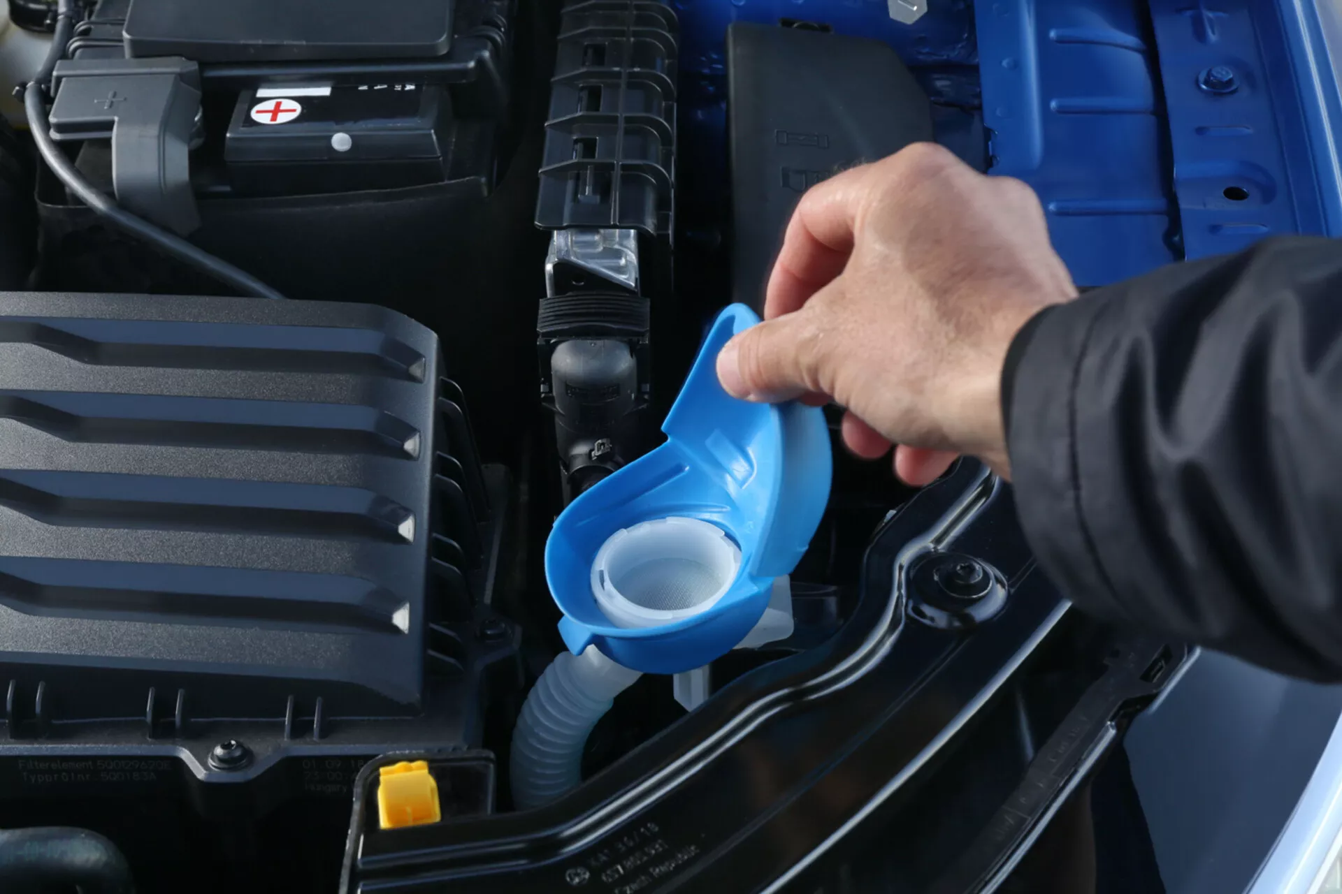 Gros plan d'une personne versant du liquide lave-glace dans le compartiment moteur d'une Skoda Scala 2019, mettant en évidence le compartiment propre et bien entretenu avec une batterie visible et des réservoirs de liquide