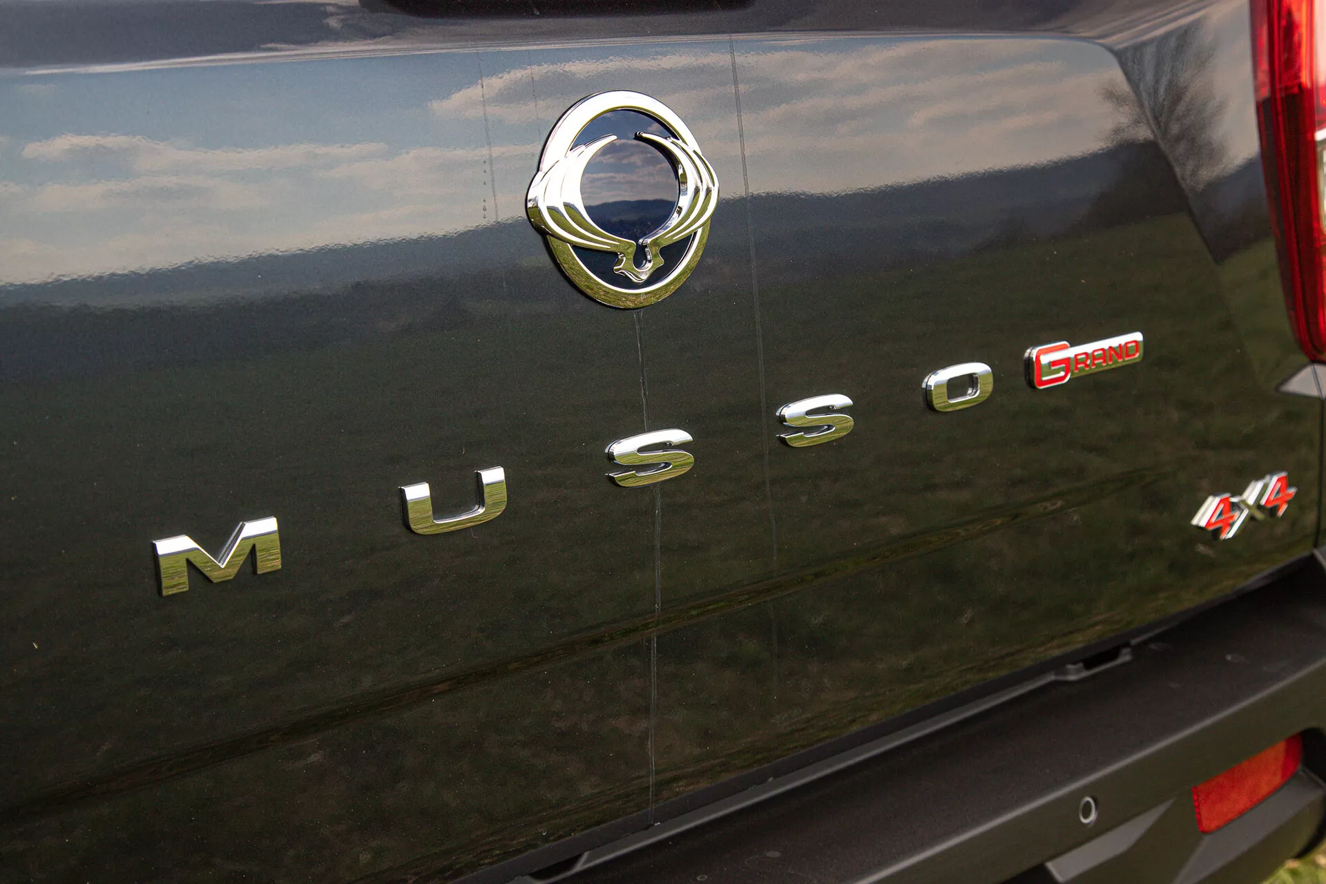 Vue rapprochée de l'arrière d'un SsangYong Musso Grand de 2022 montrant l’inscription chromée ‘MUSSO’, l’emblème argenté du fabricant avec un motif stylisé d'oiseau, et le badge rouge ‘E-XDI Sport’ indiquant le type de moteur et le niveau de performance, mis contre l'extérieur arrière sombre, possiblement noir, du véhicule avec des détails chromés réfléchissants et texture.