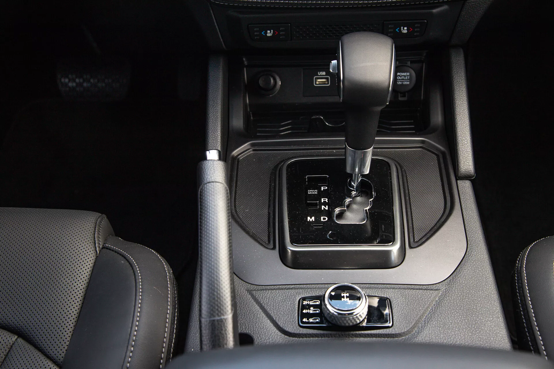 Vue détaillée de la console centrale du SsangYong Musso 2022 avec levier de vitesse automatique, frein de stationnement électronique, bouton de démarrage/arrêt, bascule du système de démarrage/arrêt automatique, porte-gobelets, prises d'entrée auxiliaires, et sièges en cuir visibles, soulignant le design moderne et les caractéristiques de luxe