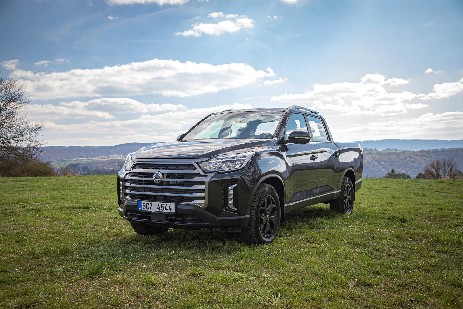 Pick-up SsangYong Musso Grand 2022 garé sur un paysage herbeux sous un ciel nuageux, exposant sa conception robuste et sa capacité tout-terrain avec une vue de trois quarts avant pendant la journée.