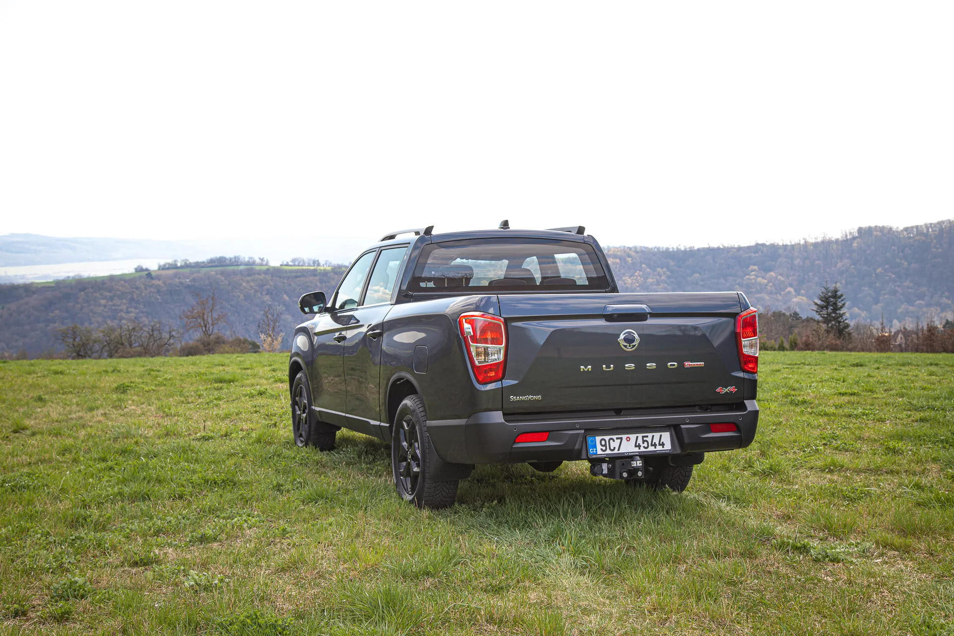 Pick-up SsangYong Musso Grand 2022 capturé sous un angle arrière mettant en lumière son apparence sportive utilitaire, sa conception à quatre portes, et sa polyvalence en extérieur contre un arrière-plan pittoresque de collines et potentiellement une rivière en journée